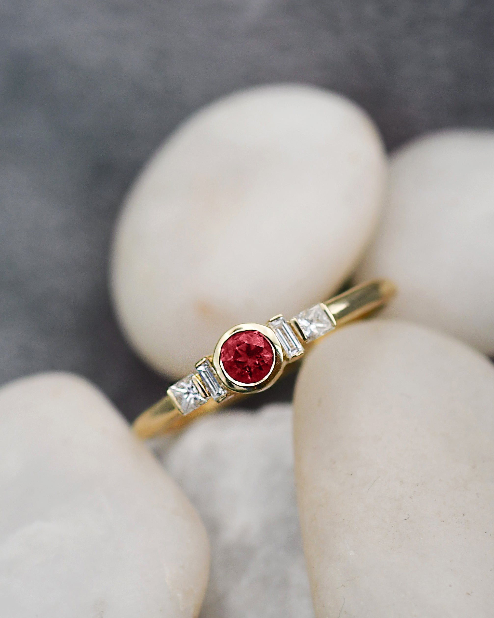 A yellow gold ring featuring a round-cut ruby gemstone in the center, flanked by two small baguette-cut clear stones, resting on smooth white stones against a dark gray background.