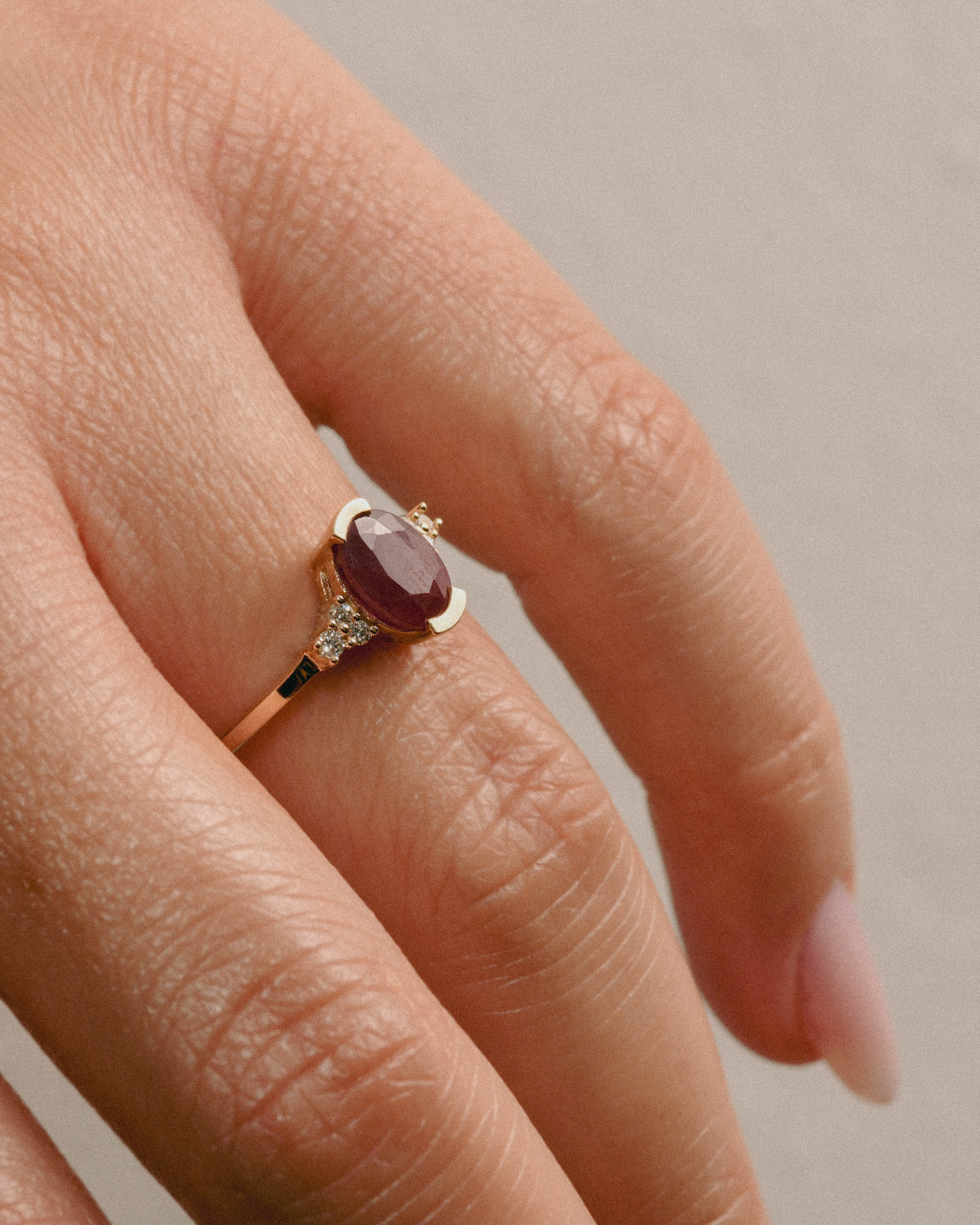 A close-up image of a person's hand showcasing a gold ring with a large natural ruby gem and smaller white diamonds.