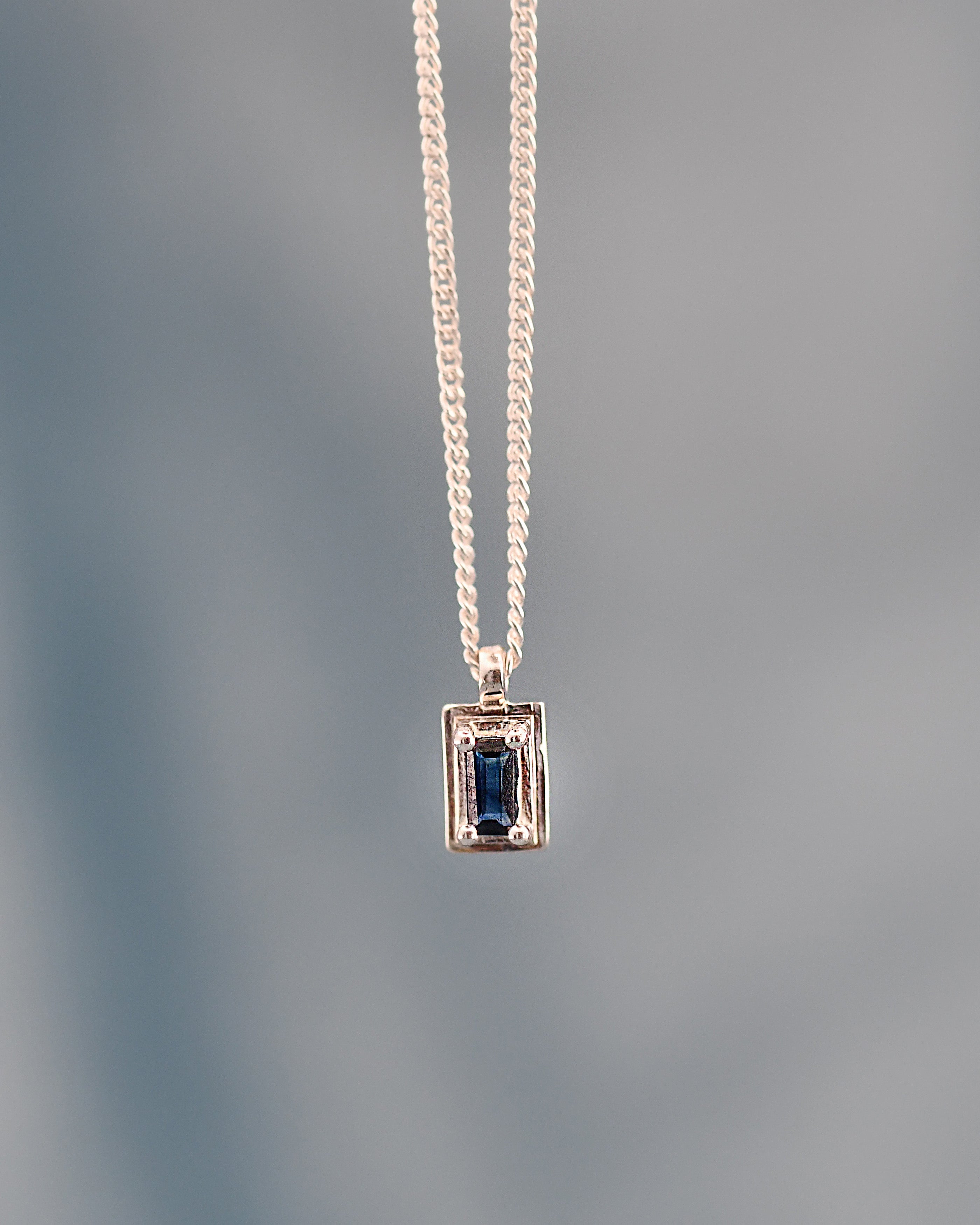 Gold necklace with a rectangular pendant featuring a blue gemstone on a gray background