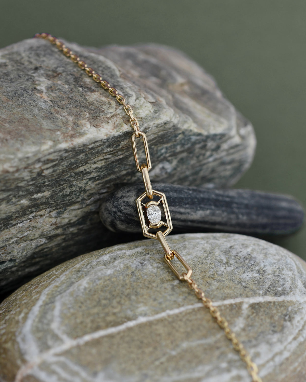 A delicate gold chain bracelet with a geometric link design and a small central clear diamond, displayed draped over stacked gray stones against a solid gray background.