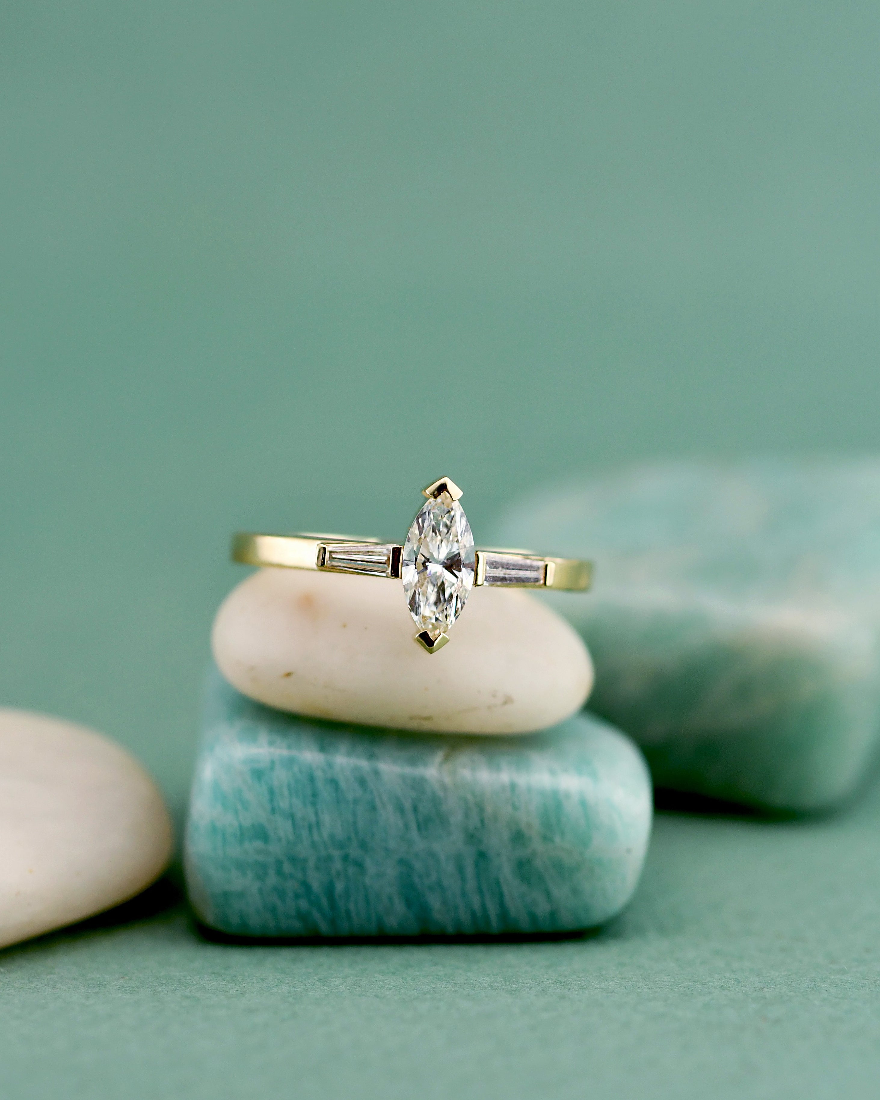 A gold ring with a marquise-cut center diamond and teiper side stones, displayed on stacked smooth stones against a soft teal-green background.