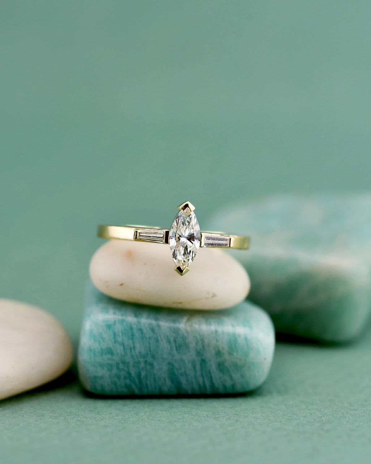 A gold ring with a marquise-cut center diamond and teiper side stones, displayed on stacked smooth stones against a soft teal-green background.