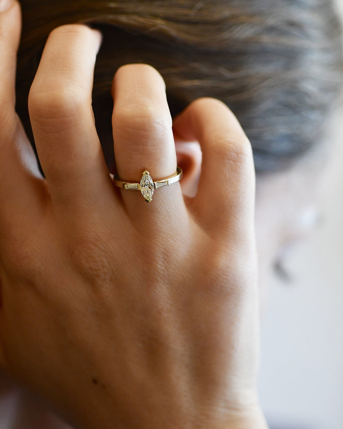 A close-up of a person's hand wearing a gold ring with a marquise-cut clear diamond on the ring finger, with dark hair in the background.