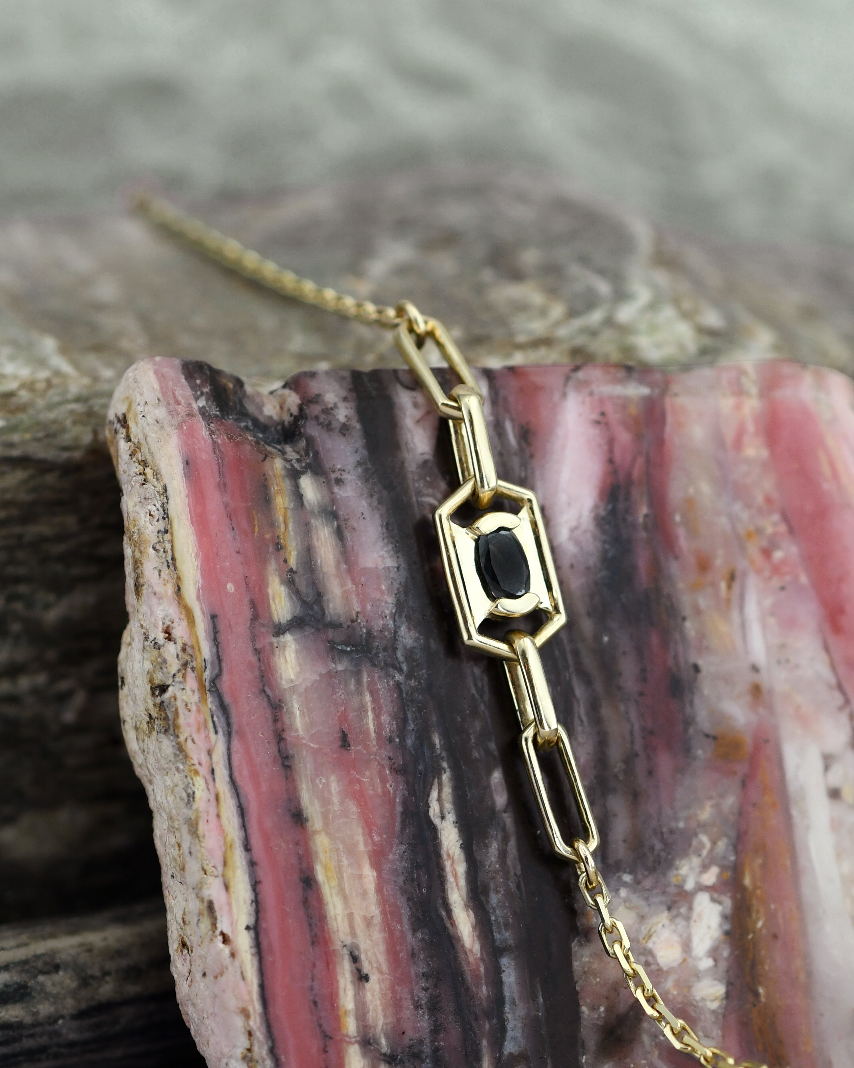 A gold chain bracelet with a geometric link pendant featuring a central black oval diamond, displayed on a pink and black banded agate rock.