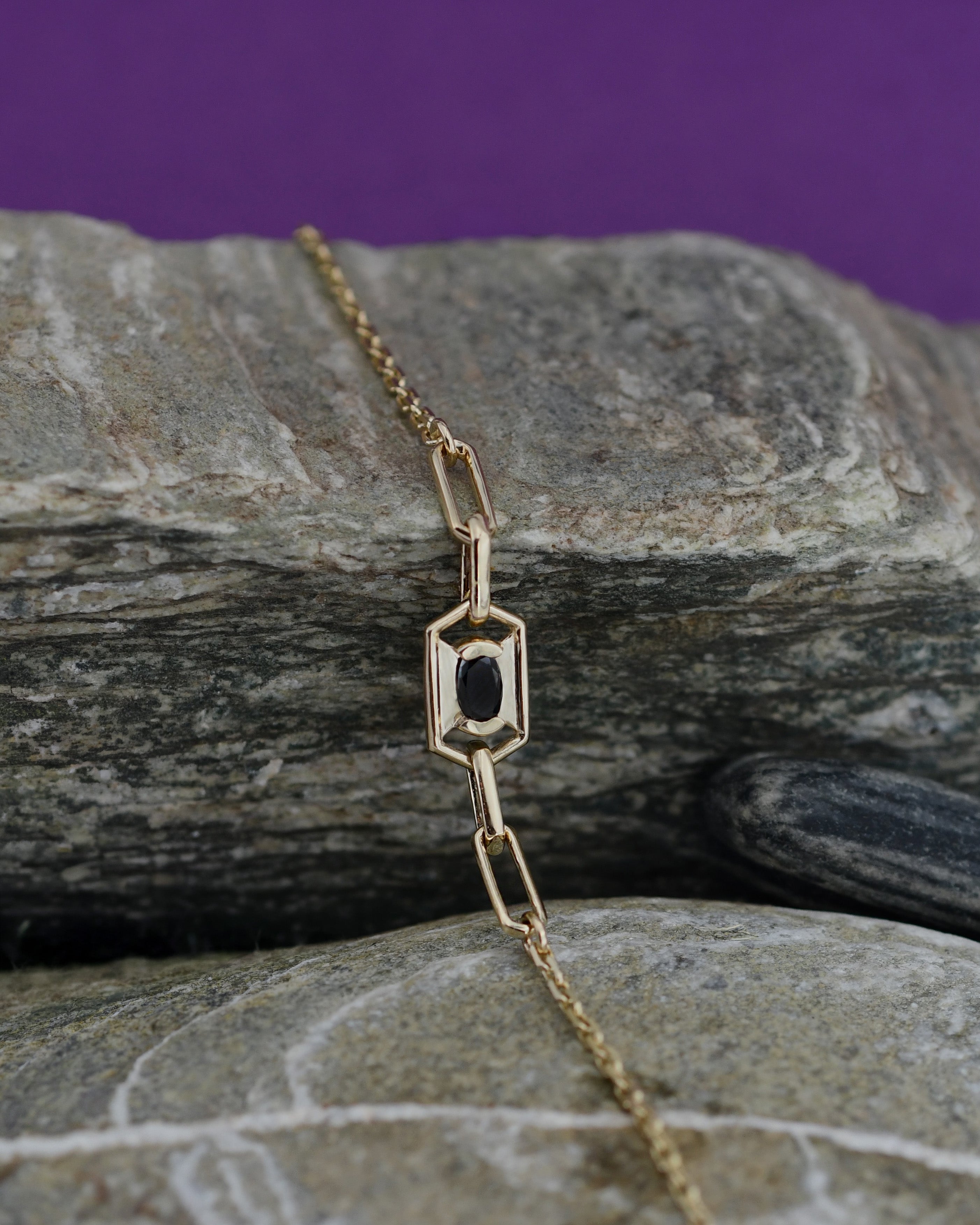 A gold chain bracelet with a geometric link pendant featuring a central black oval diamond, displayed on stones.