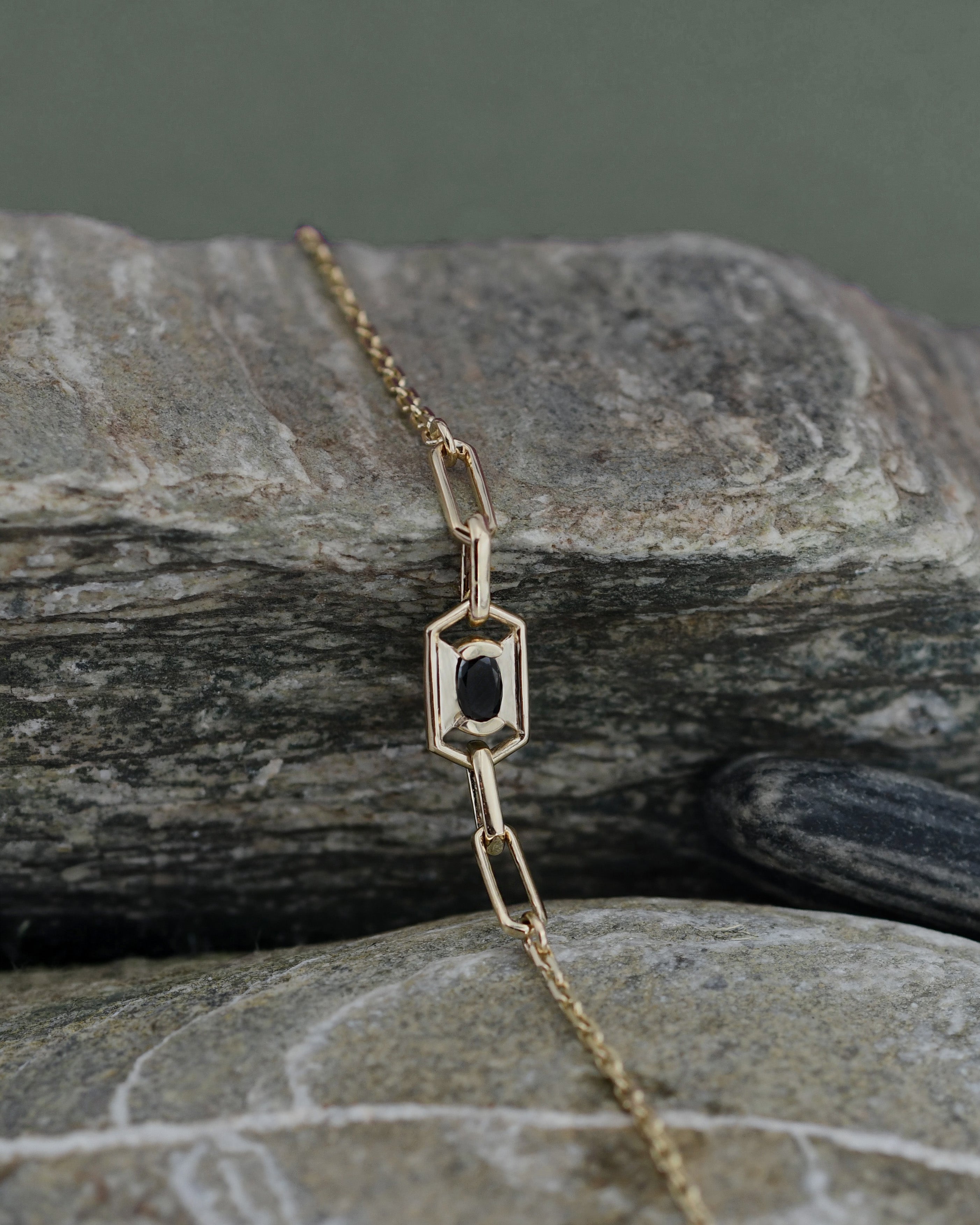 A gold chain bracelet with a geometric link pendant featuring a central black oval diamond, displayed on stones.