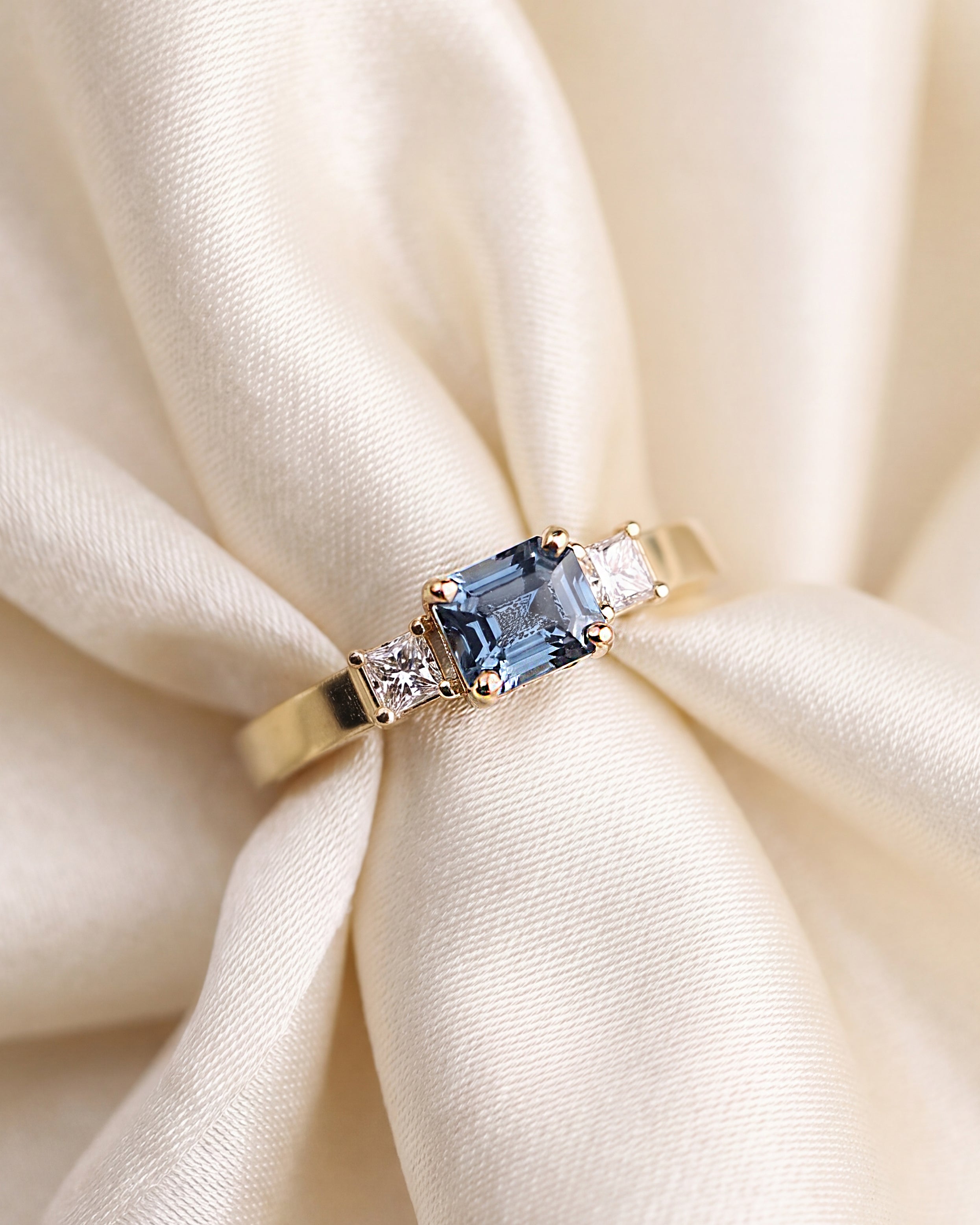 A gold three-stone ring featuring a central emerald-cut light blue gemstone and two round clear side diamonds, displayed on a metallic cylindrical stand against a soft gray background.