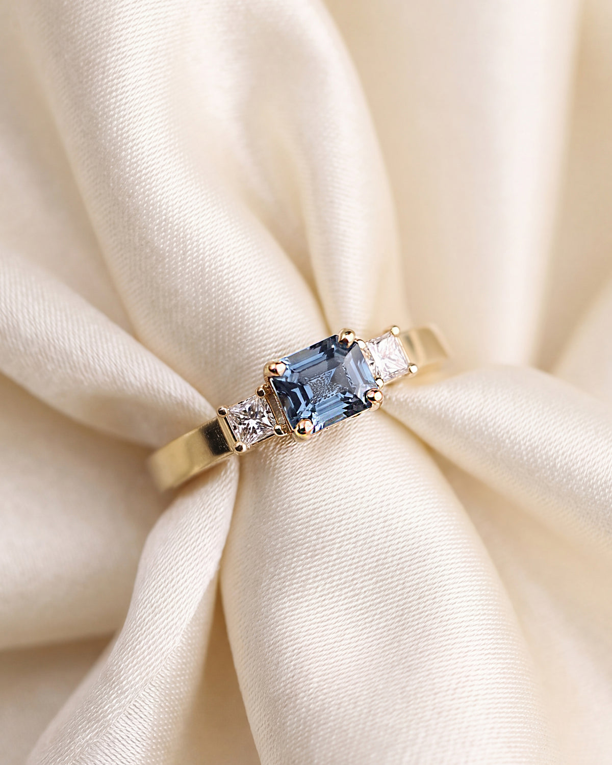 A gold three-stone ring featuring a central emerald-cut light blue gemstone and two round clear side diamonds, displayed on a metallic cylindrical stand against a soft gray background.