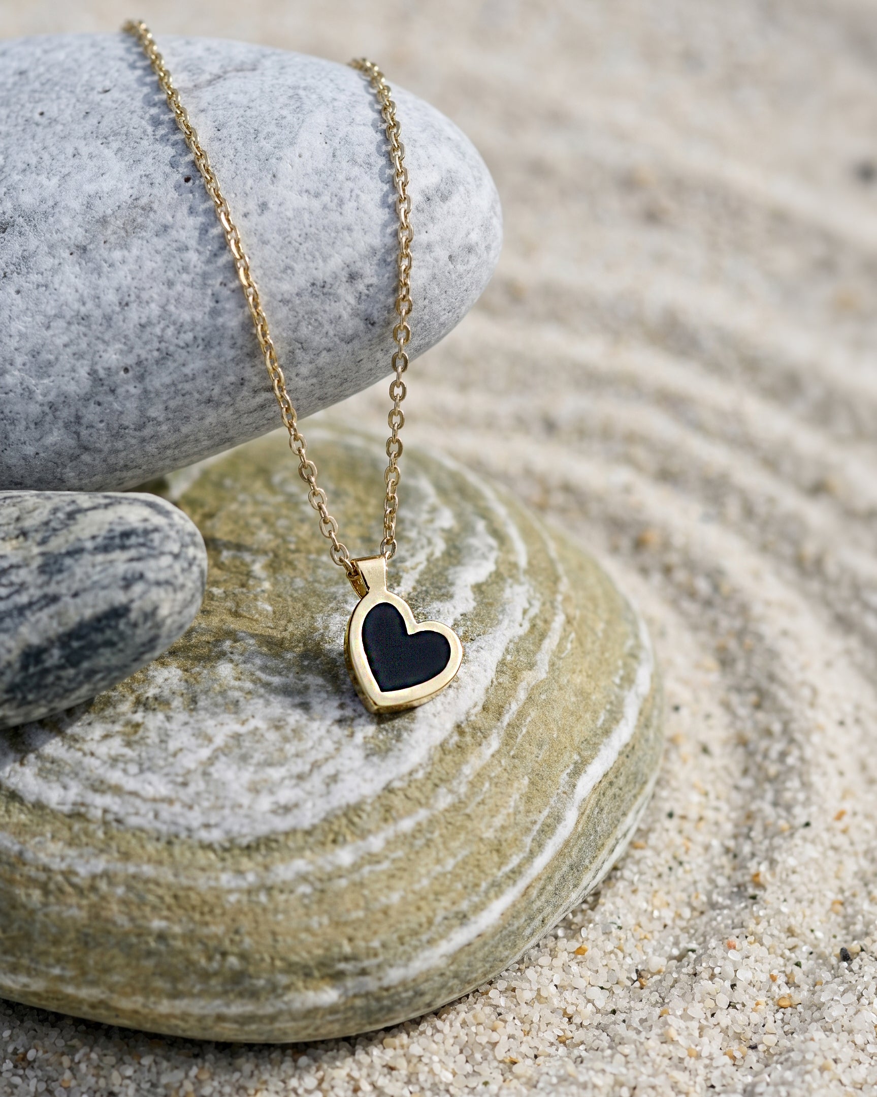 A gold heart pendant necklace with a black inlay, resting on stacked gray and green stones over a bed of sand.