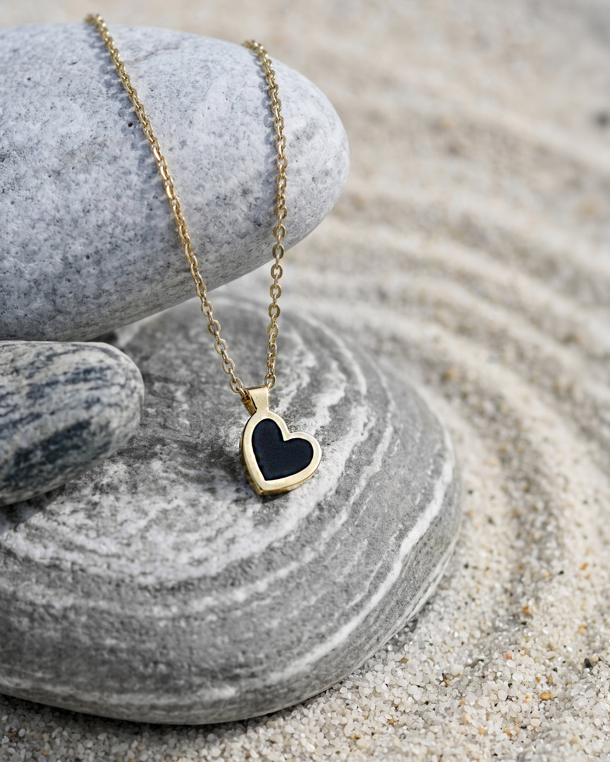 A gold heart pendant necklace with a black inlay, resting on stacked gray and green stones over a bed of sand.