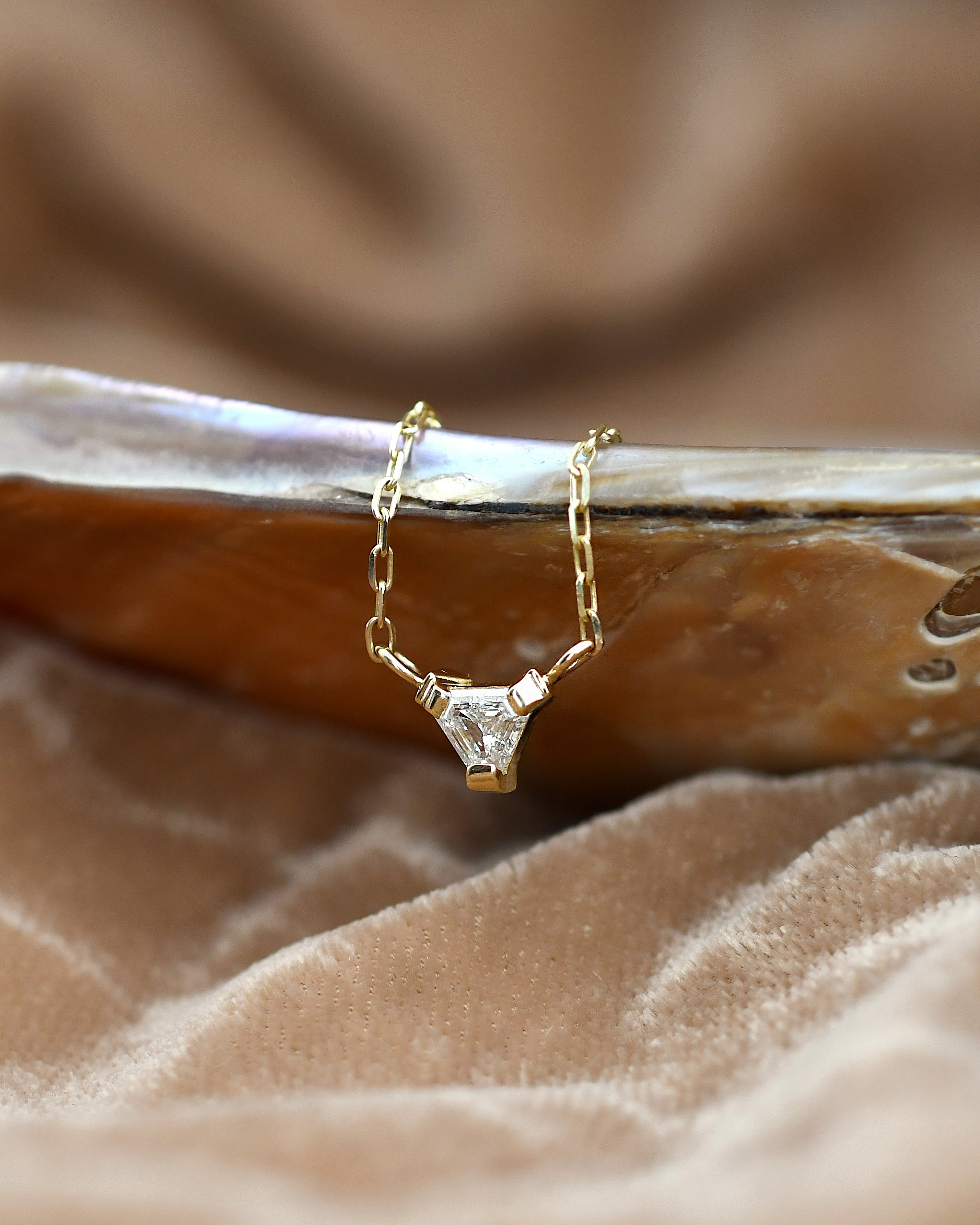 A delicate gold chain necklace with a geometric clear white diamond pendant, resting on a polished shell over a soft beige fabric background.