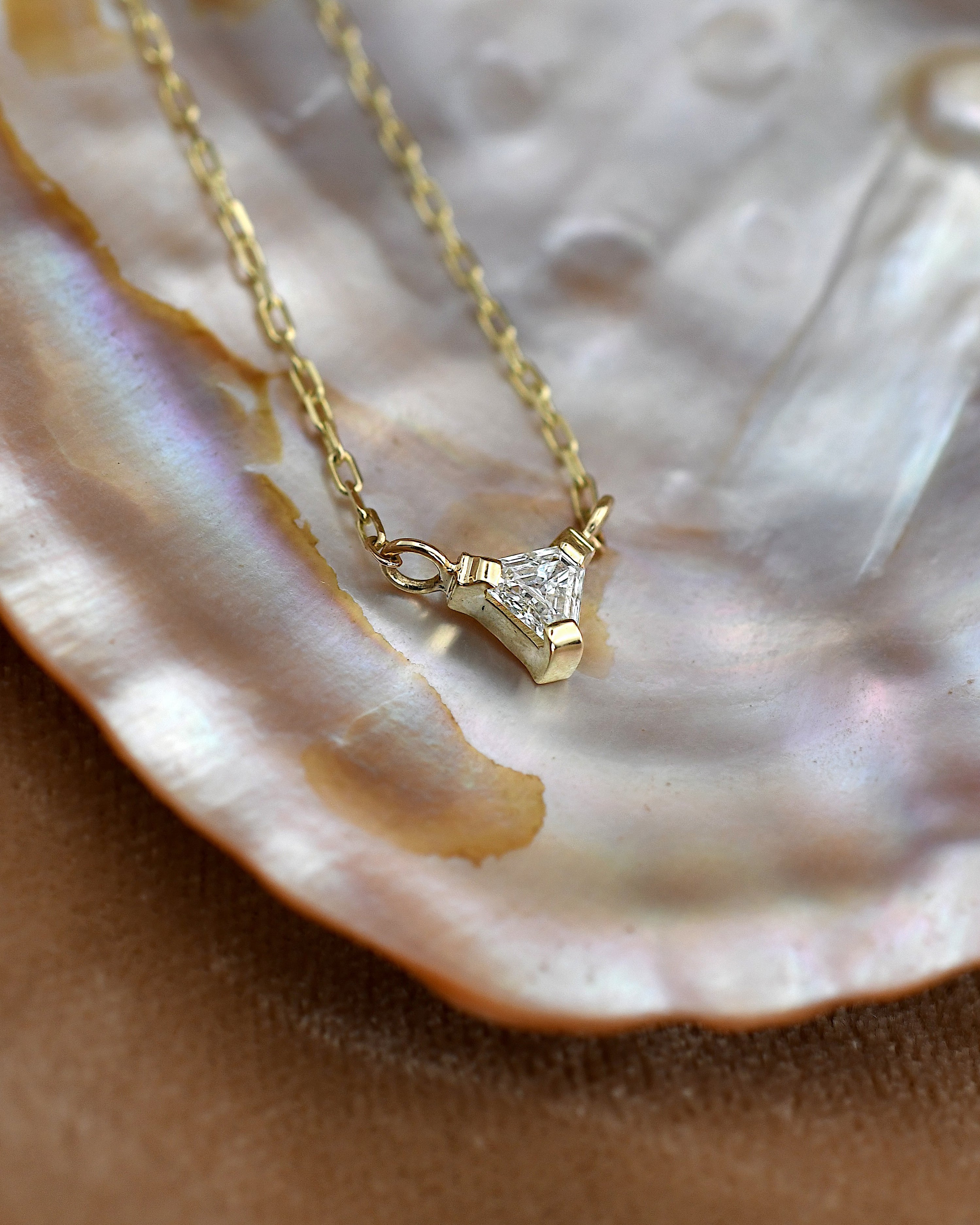 A gold chain necklace with a small geometric triangular pendant set with a clear white diamond, displayed on a glossy iridescent shell over a brown surface.