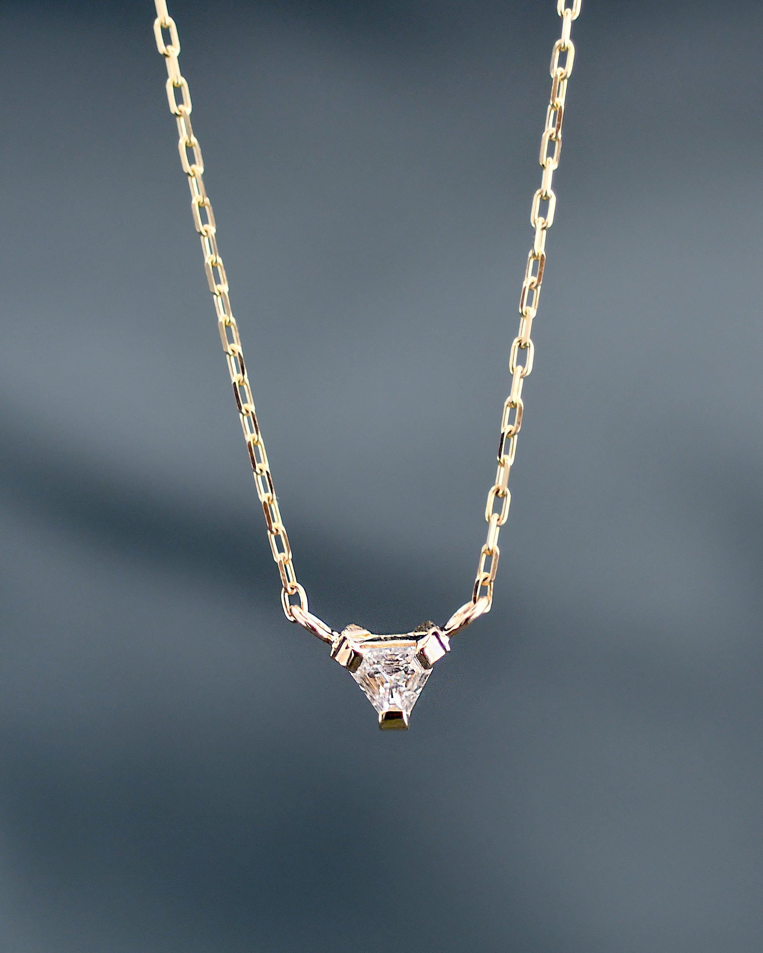 A delicate gold chain necklace with a small triangular pendant set with a clear white diamond, shown against a dark gray gradient background.