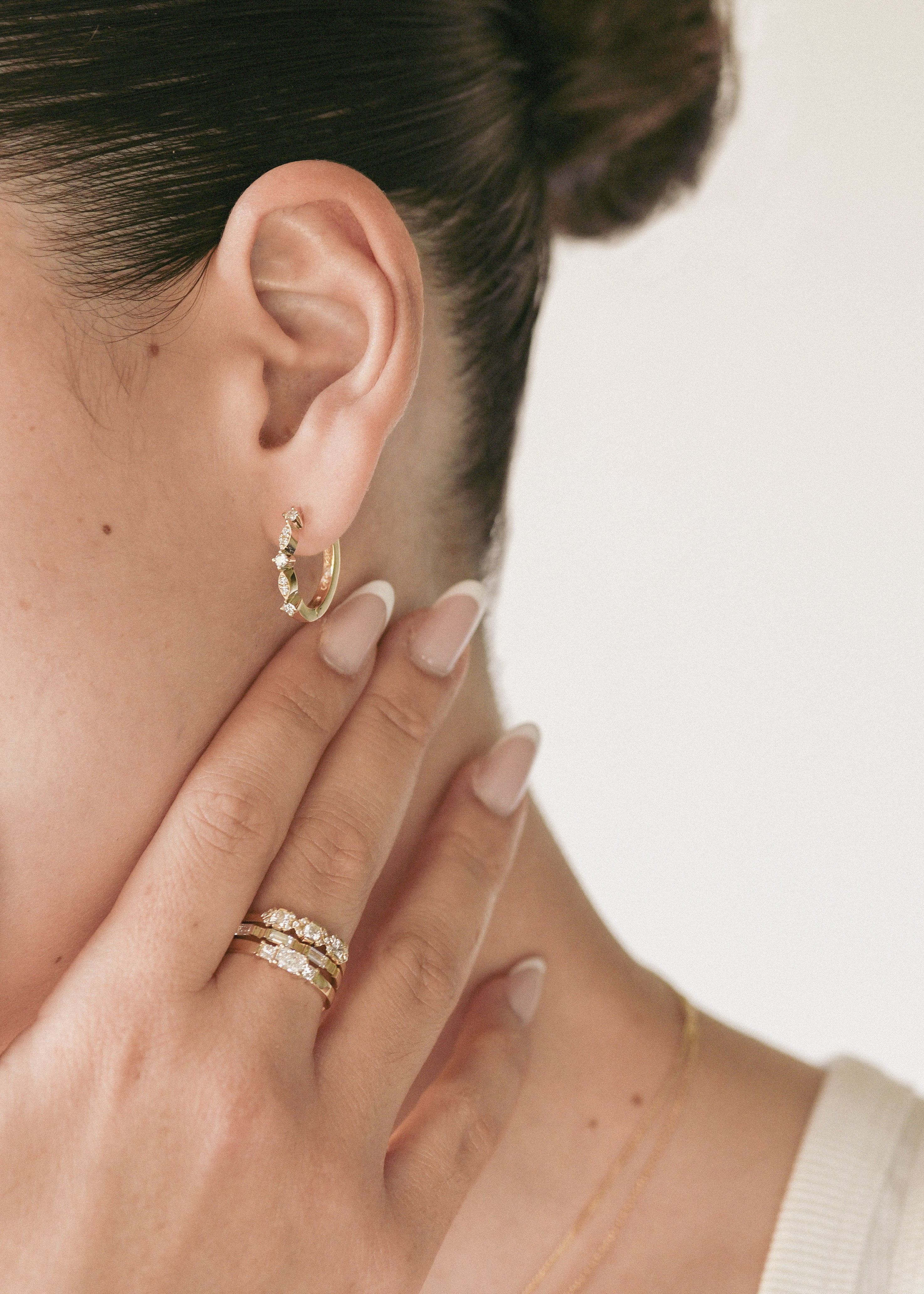 A close-up lifestyle shot of a model wearing a delicate gold crescent moon hoop earring on her ear, with her hand resting against her cheek. She is also wearing two gold rings on her finger.