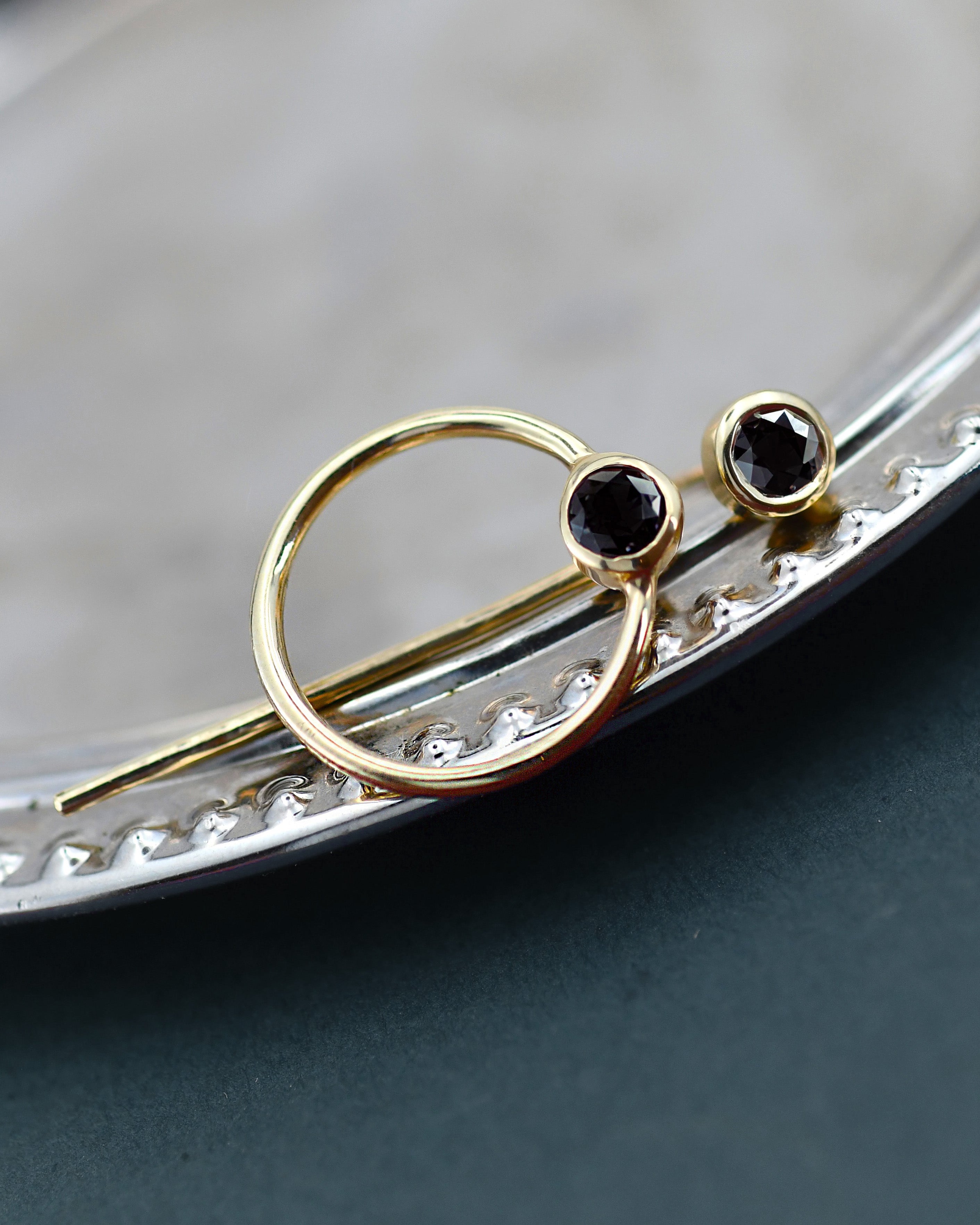 A pair of gold stud earrings with round black faceted gemstones, resting on the edge of a silver tray over a dark surface.
