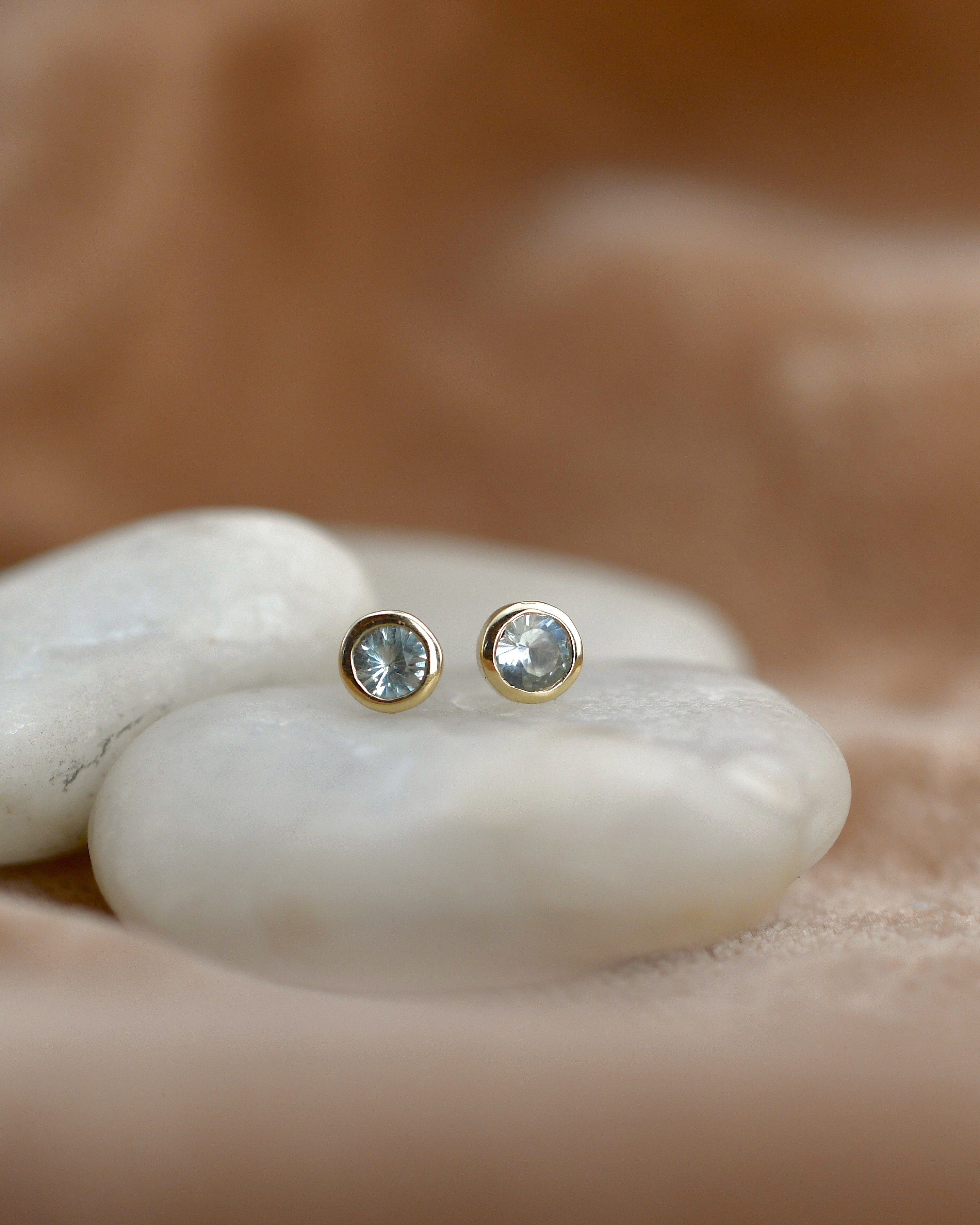 A pair of gold stud earrings with round clear gemstones resting on smooth white stones against a warm beige background.