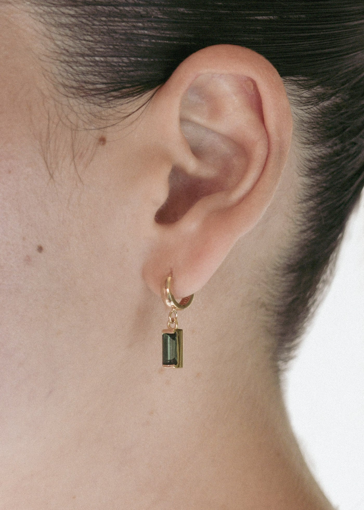 A close-up of a person's ear wearing a small gold huggie hoop earring with a rectangular dark green gemstone dangle.