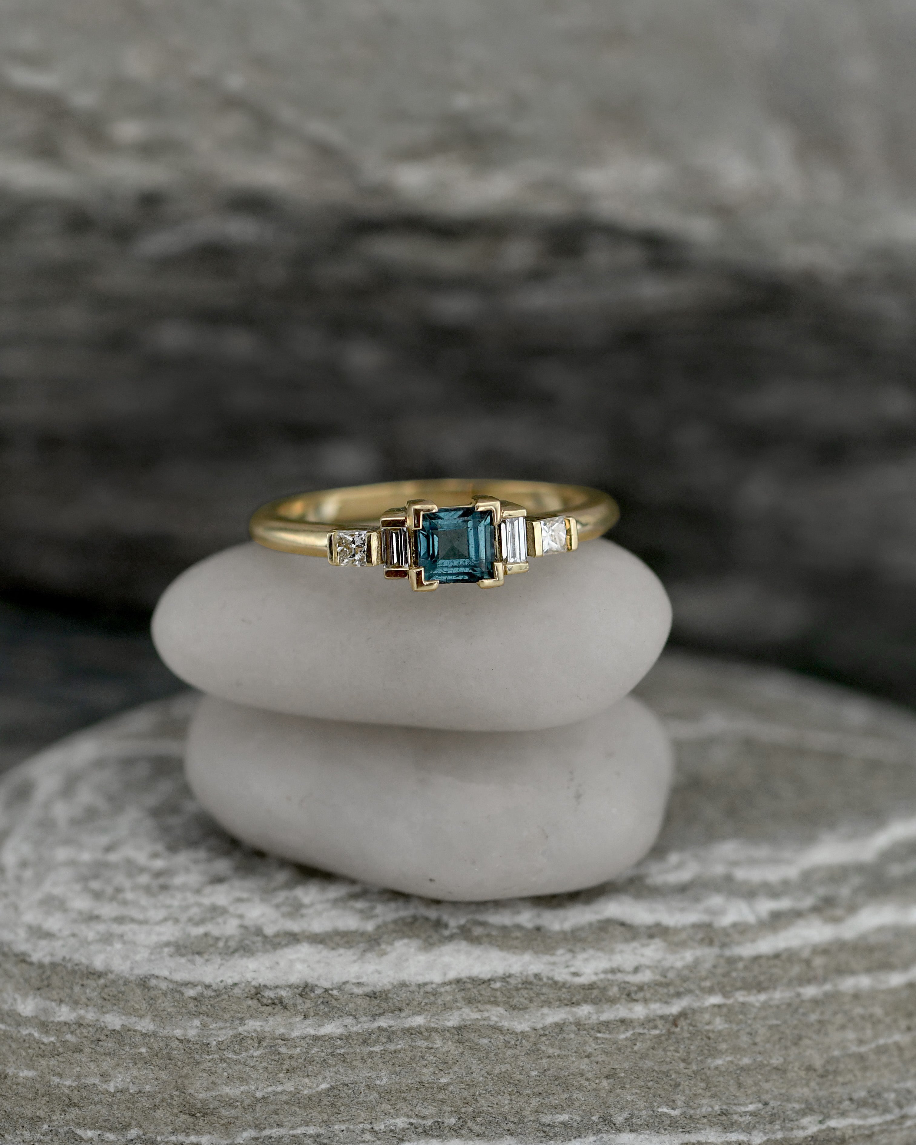 A gold ring with a central blue tourmaline gemstone and small side diamonds, displayed on stacked white stones against a blurred natural stone background.