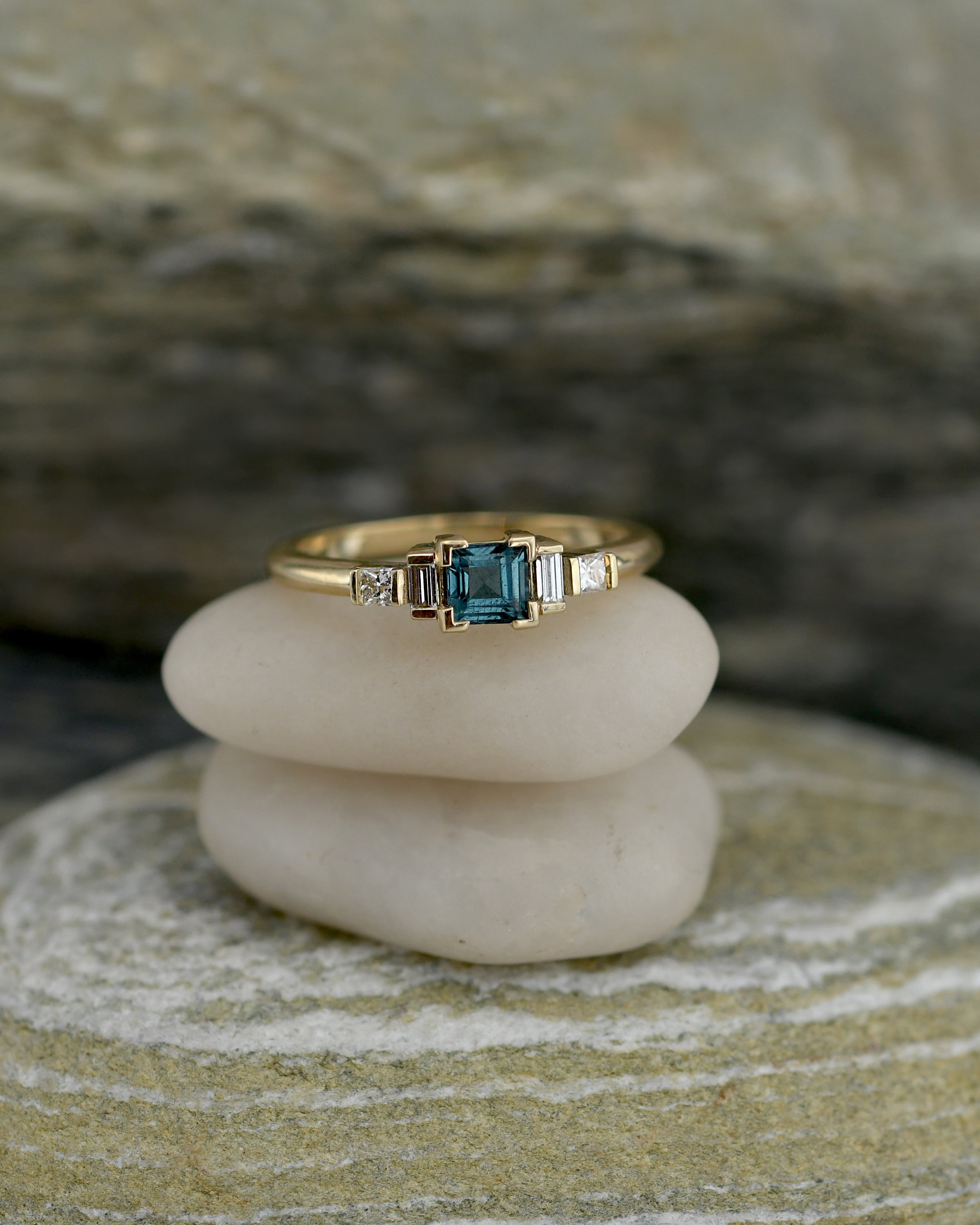 A gold ring with a central blue tourmaline gemstone and small side diamonds, displayed on stacked white stones against a blurred natural stone background.