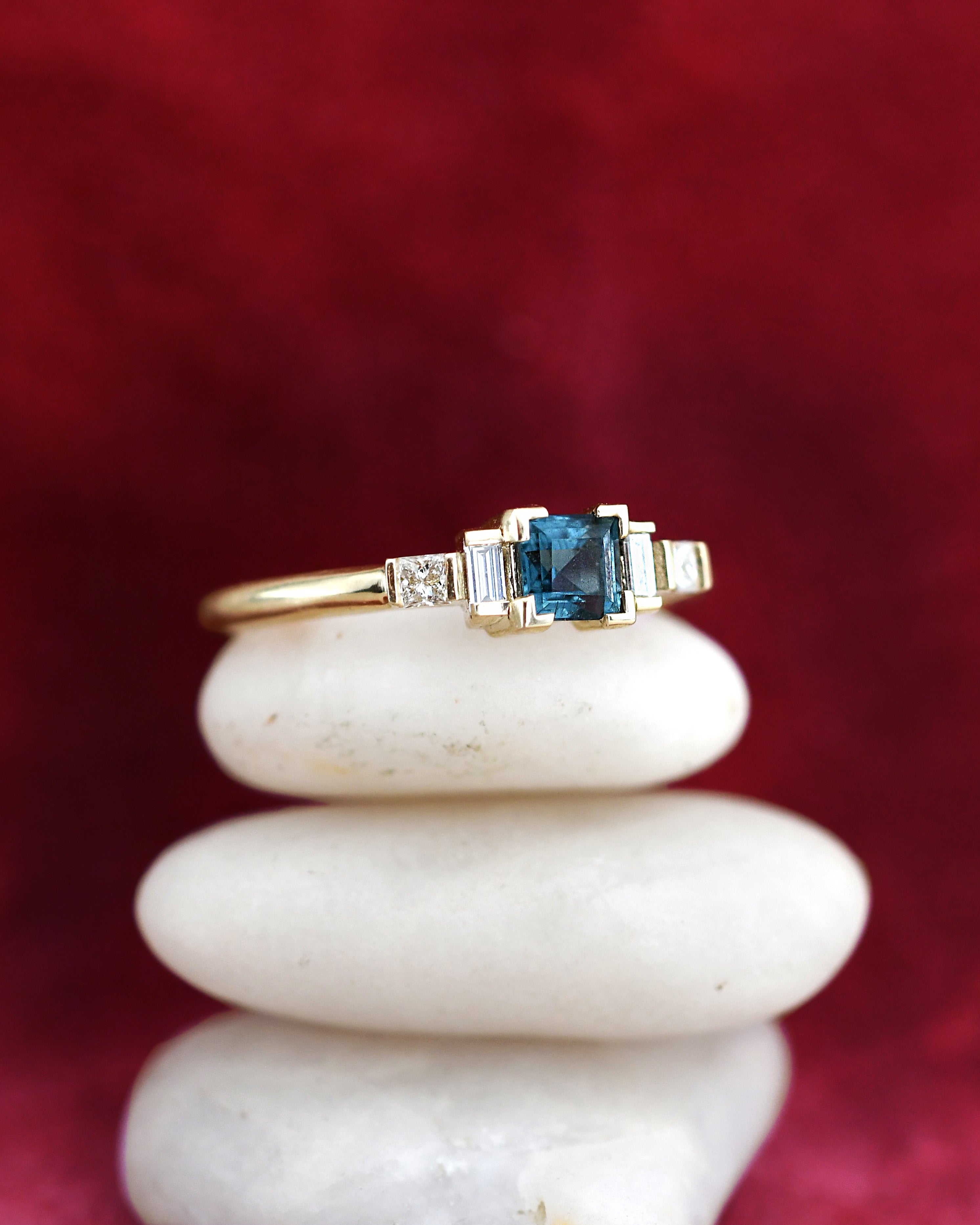 A yellow gold ring featuring a square-cut blue tourmaline center stone and small side diamonds, displayed on a stack of three white stones against a dark red velvet background.