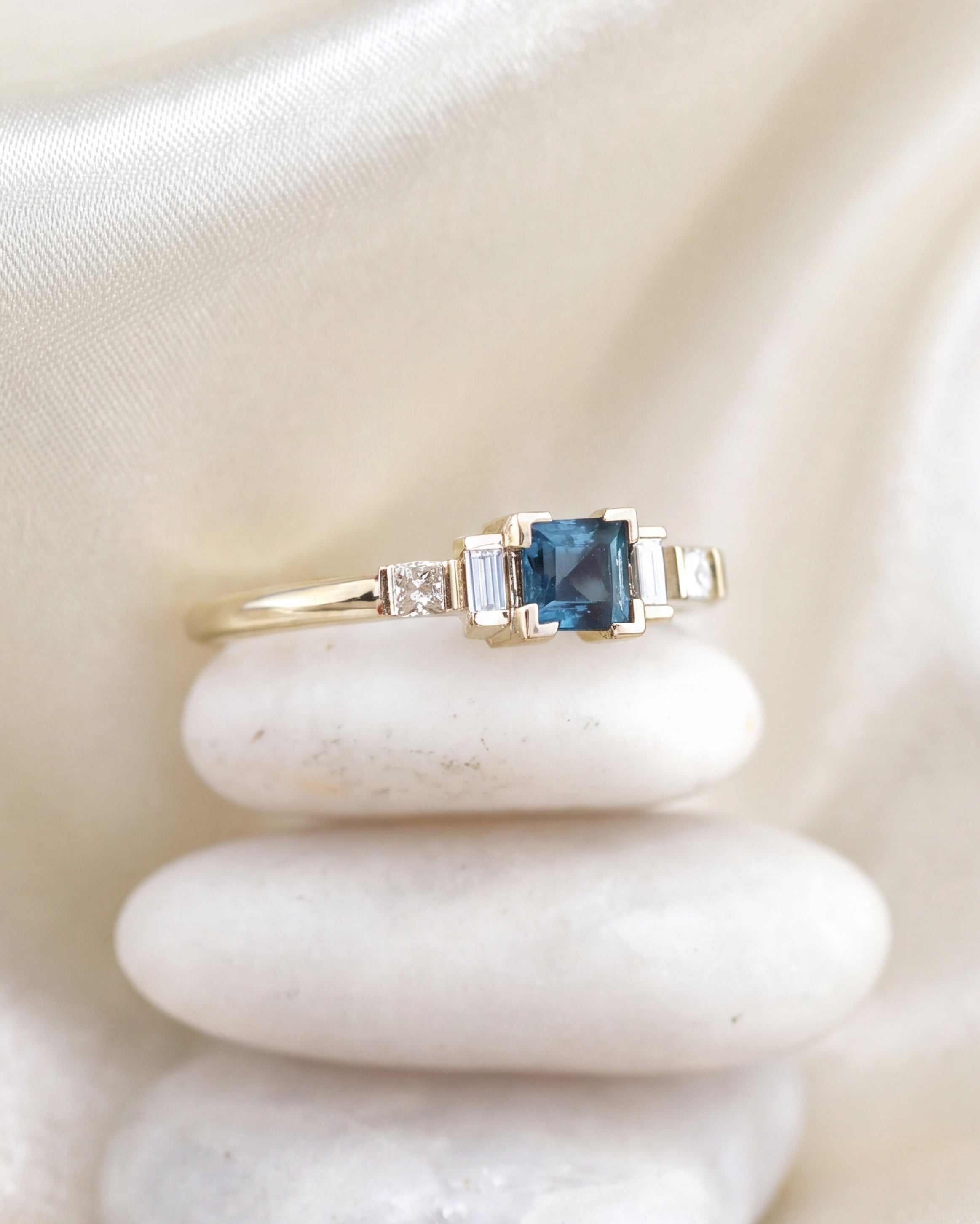A yellow gold ring featuring a square-cut blue tourmaline center stone and small side diamonds, displayed on a stack of three white stones against a dark r background.