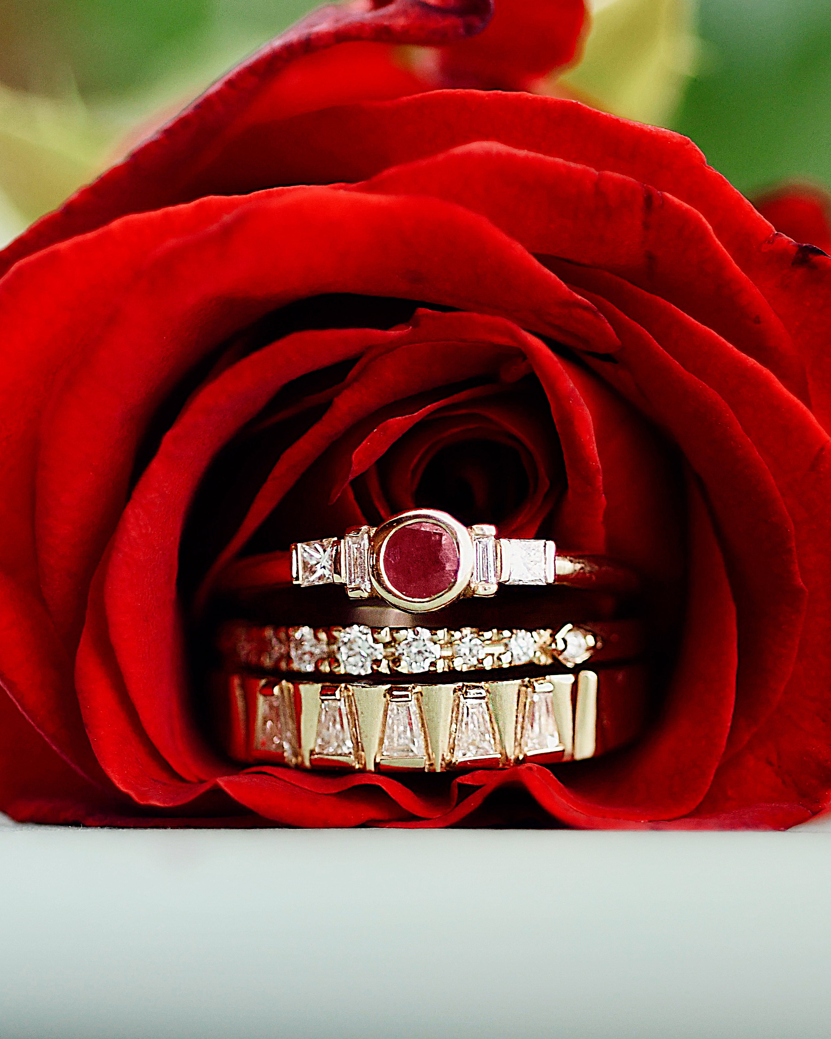 Three gold rings with diamonds and a ruby gemstone, displayed on a vibrant red rose.
