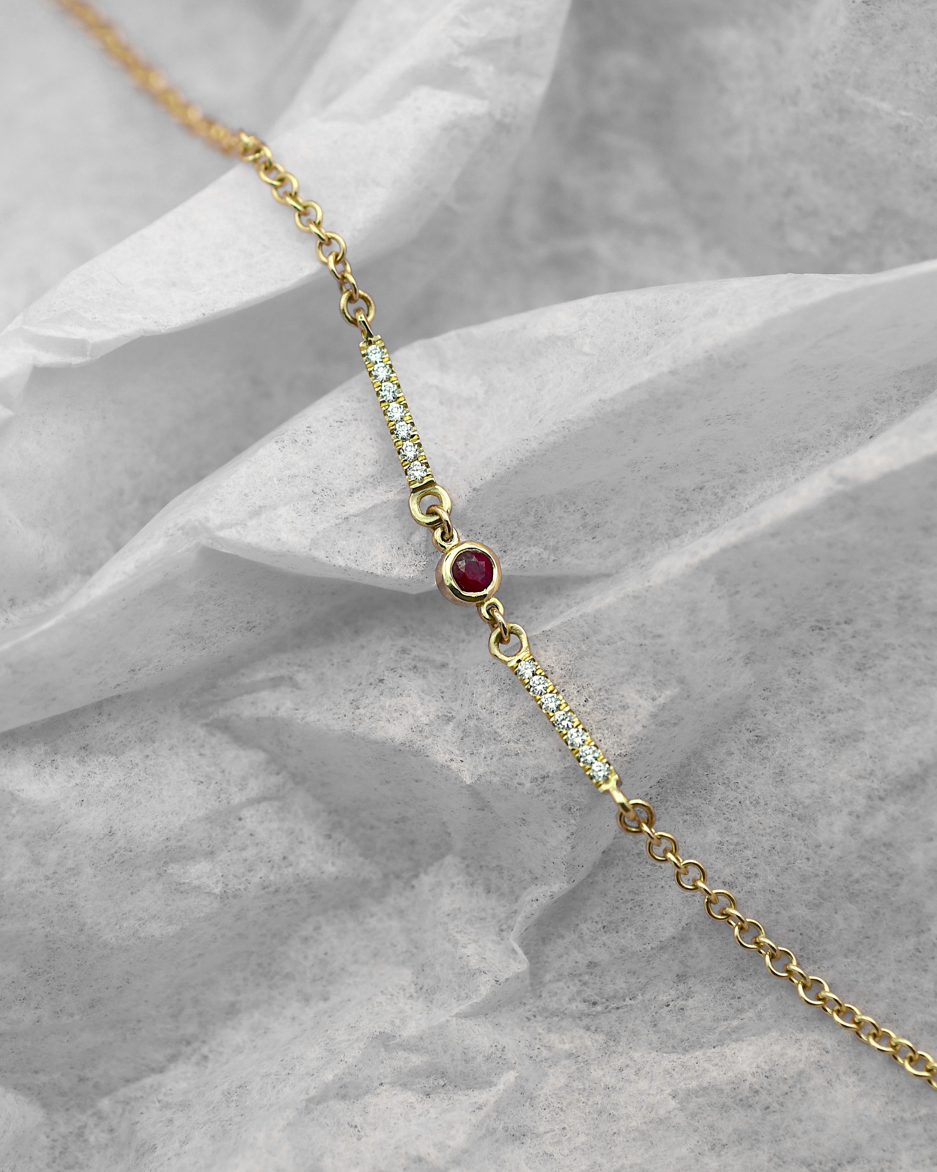 A delicate gold chain bracelet featuring a central round red ruby gemstone and two vertical rows of small clear diamonds, displayed on a crumpled gray fabric background.