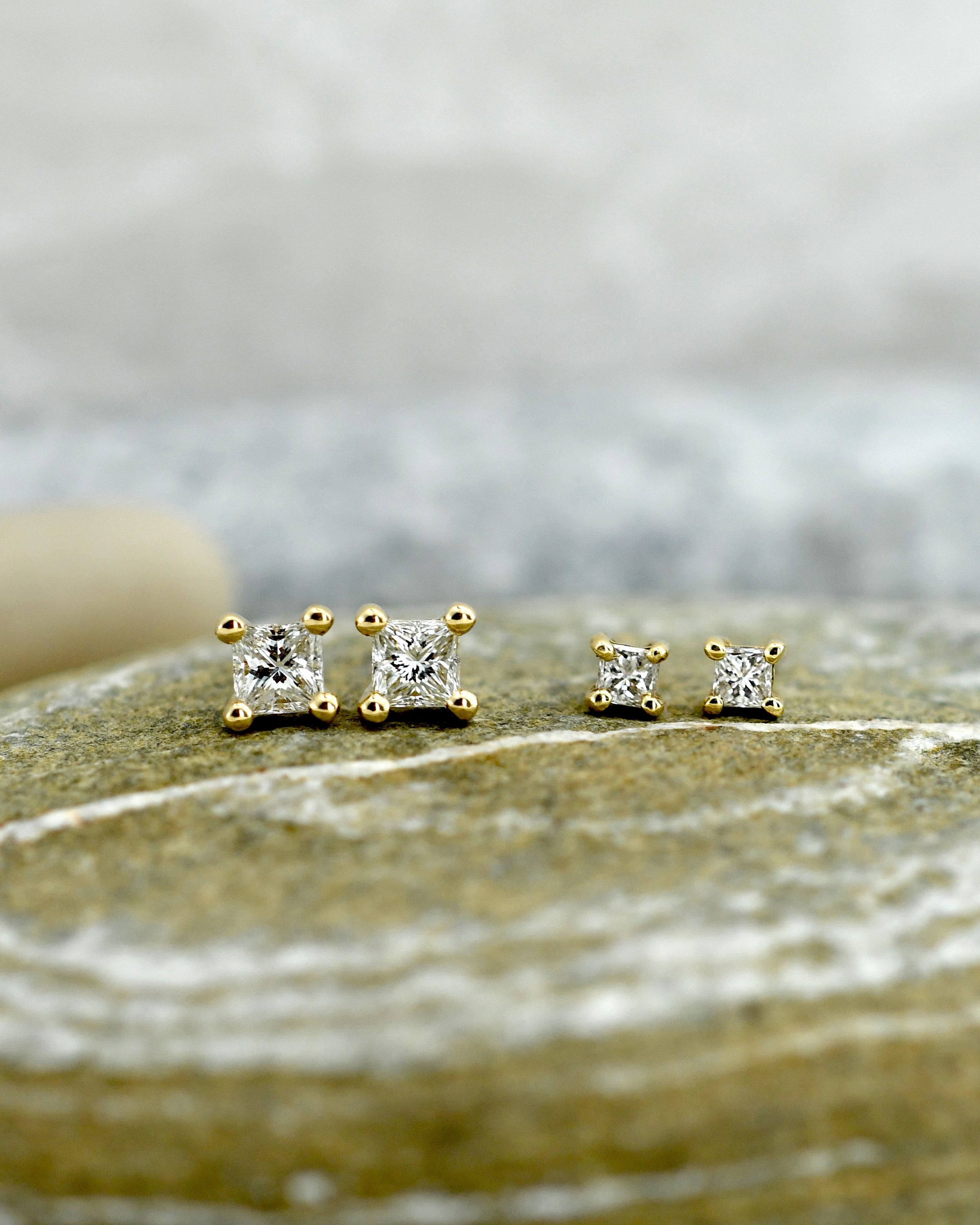 two pairs of gold stud earrings with clear princess cut diamonds in square settings, displayed on a textured stone surface with a soft blurred background.