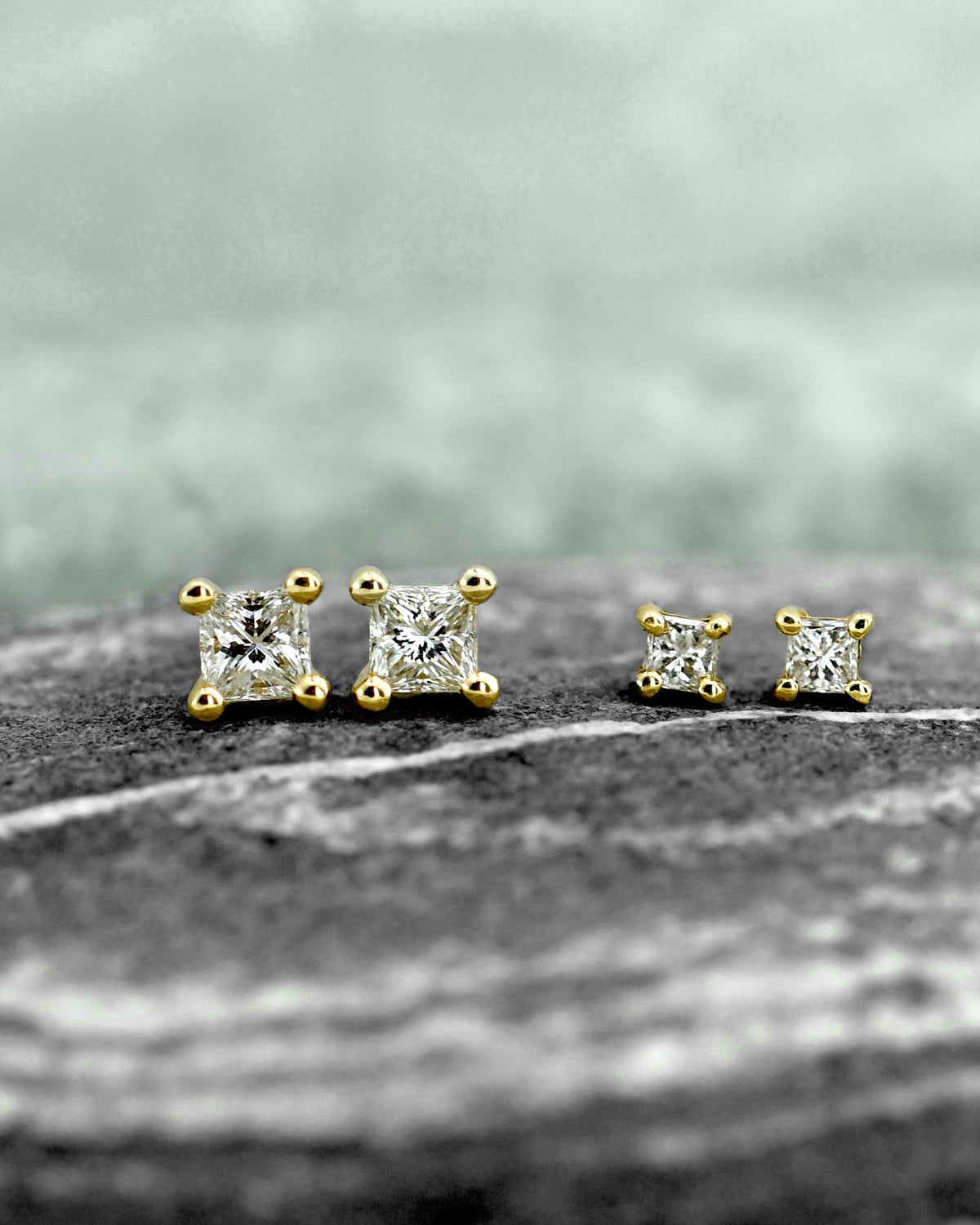 two pairs of gold stud earrings with clear princess cut diamonds in square settings, displayed on a textured stone surface with a soft blurred background.