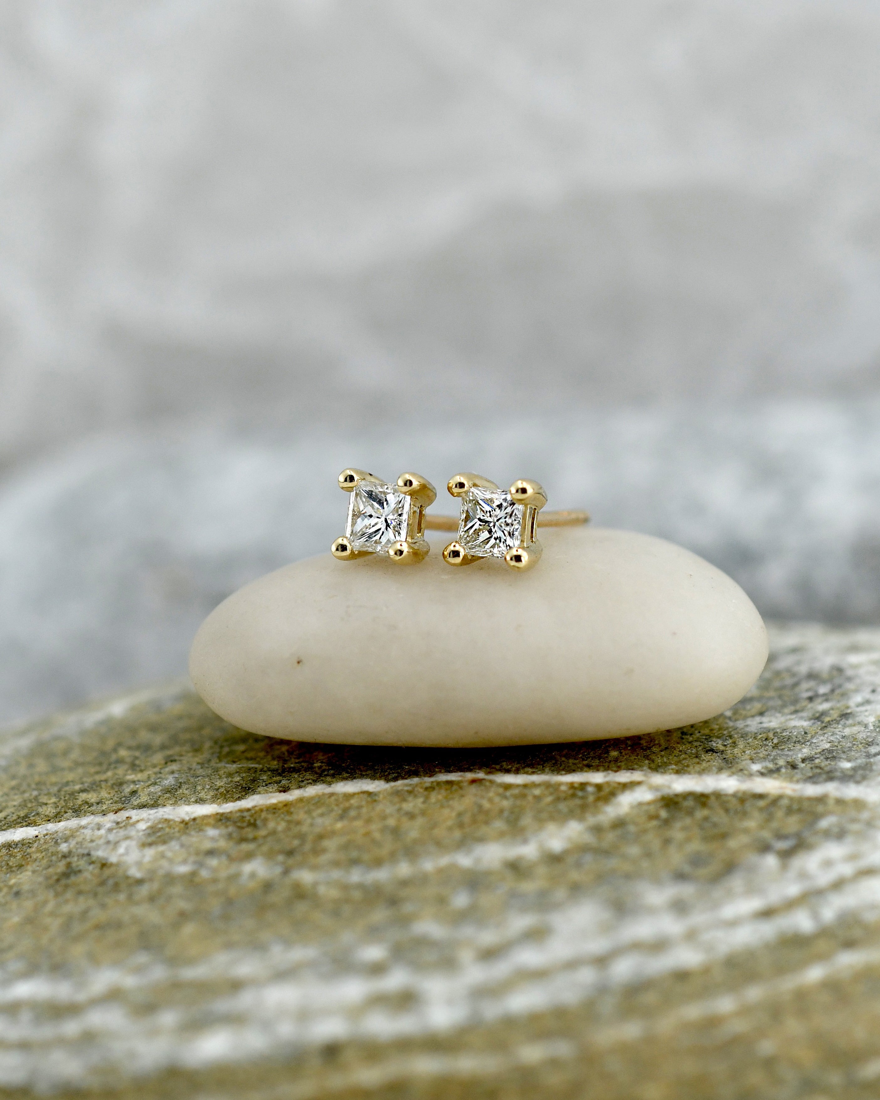 Pair of gold stud earrings with clear princess cut diamonds in square settings, displayed on a textured stone surface with a soft blurred background.