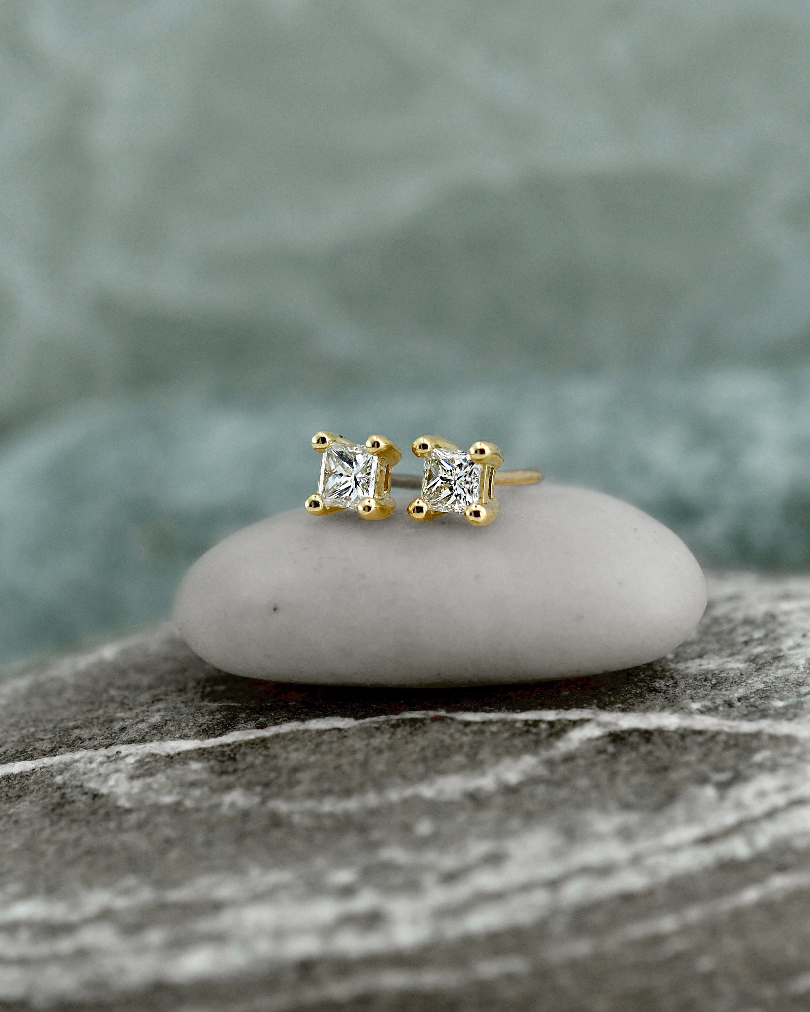 Pair of gold stud earrings with clear princess cut diamonds in square settings, displayed on a textured stone surface with a soft blurred background.
