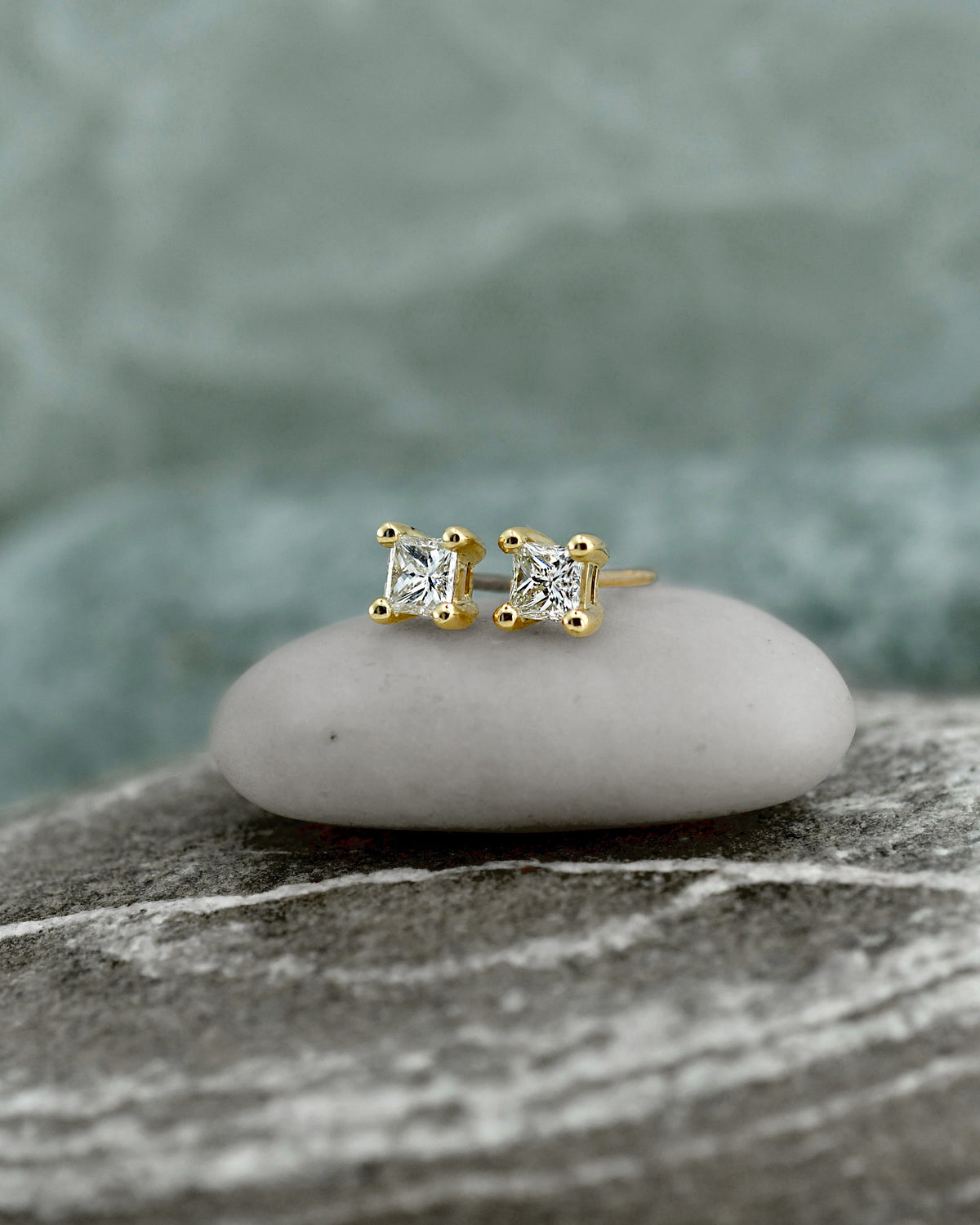 Pair of gold stud earrings with clear princess cut diamonds in square settings, displayed on a textured stone surface with a soft blurred background.