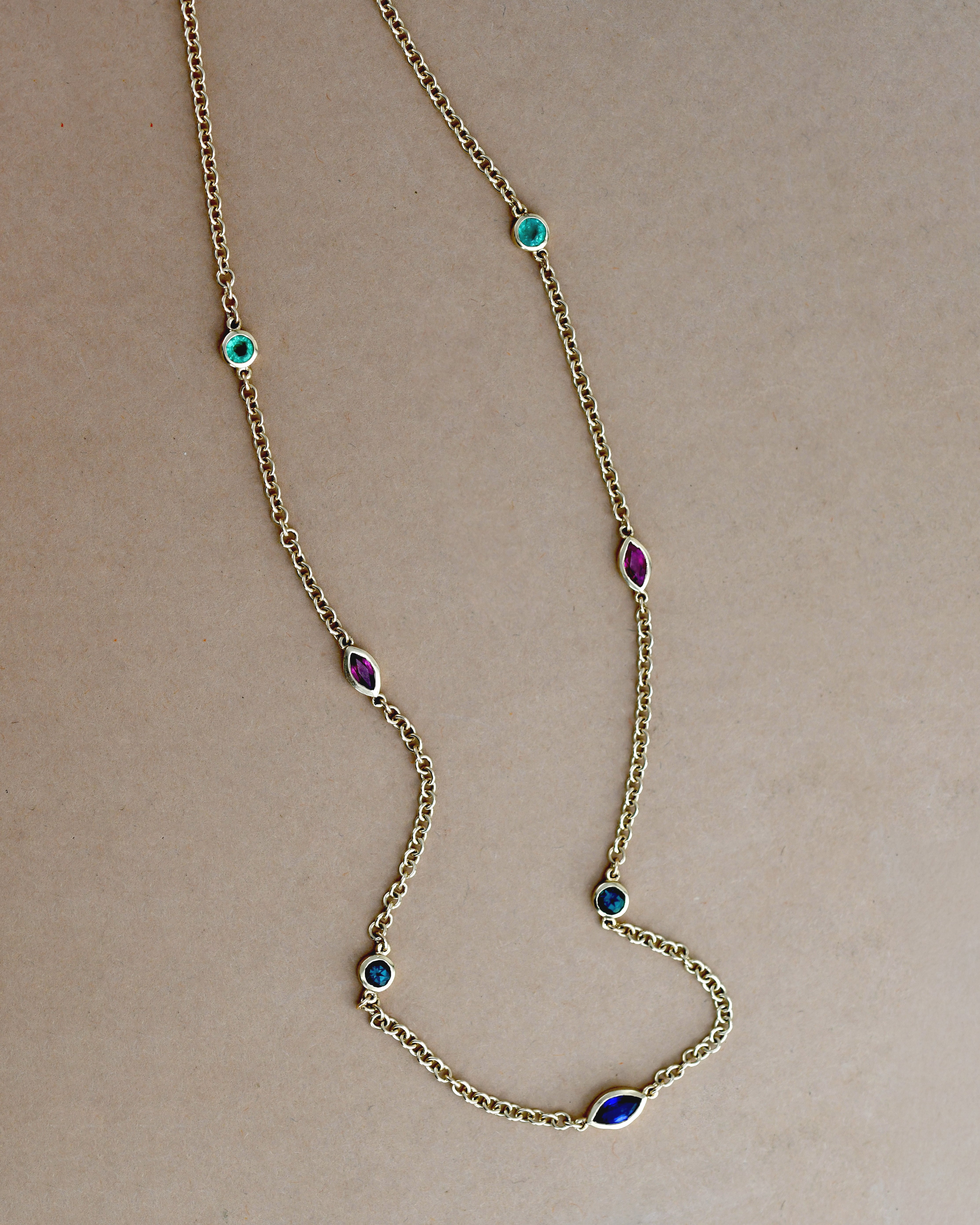 A delicate gold chain necklace featuring multiple small, colorful gemstone settings along the chain, including green round -cut emerald, red ruby marquise-cut stones, blue round -cut tourmaline, and  blue sapphire marquise-cut stones