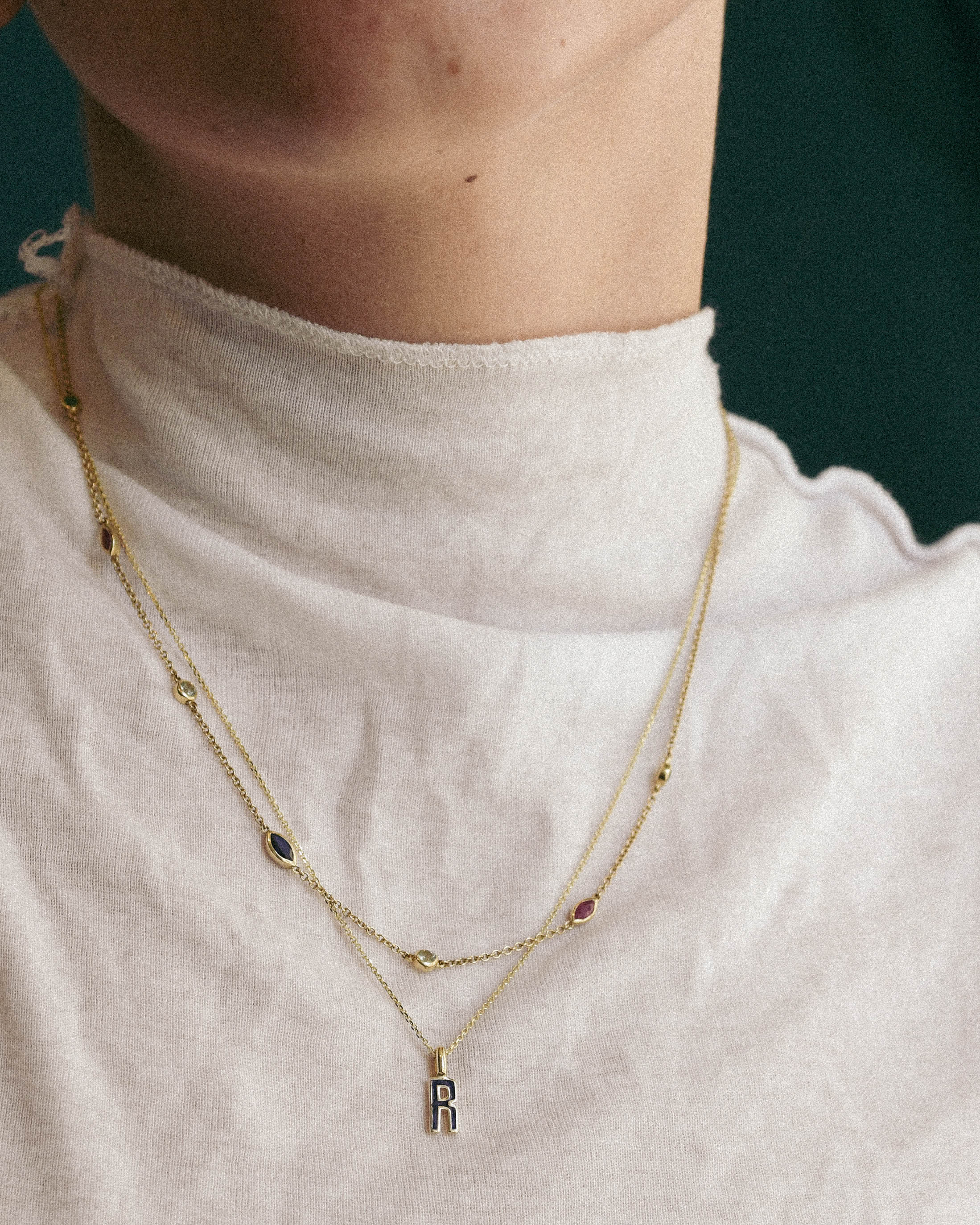 Person wearing a gold necklace with a letter 'R' pendant, and A delicate gold chain necklace featuring multiple small, colorful gemstone settings along the chain, including green round -cut emerald, red ruby marquise-cut stones, blue round -cut tourmaline, and d blue sapphire marquise-cut stones