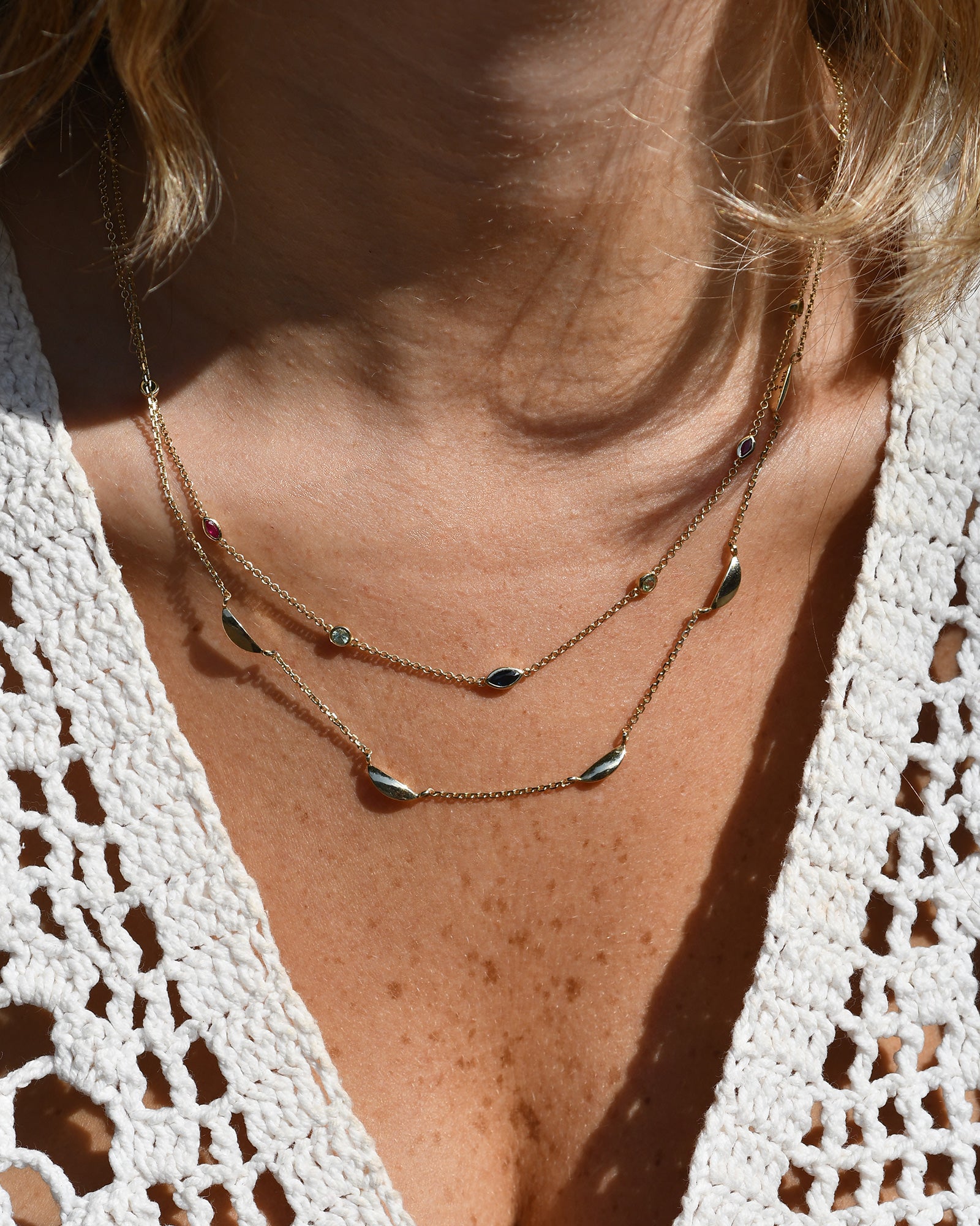 A delicate gold chain necklace featuring multiple small, colorful gemstone settings along the chain, including green round -cut emerald, red ruby marquise-cut stones, blue round -cut tourmaline, and d blue sapphire marquise-cut stones, displayed on a woman's neck