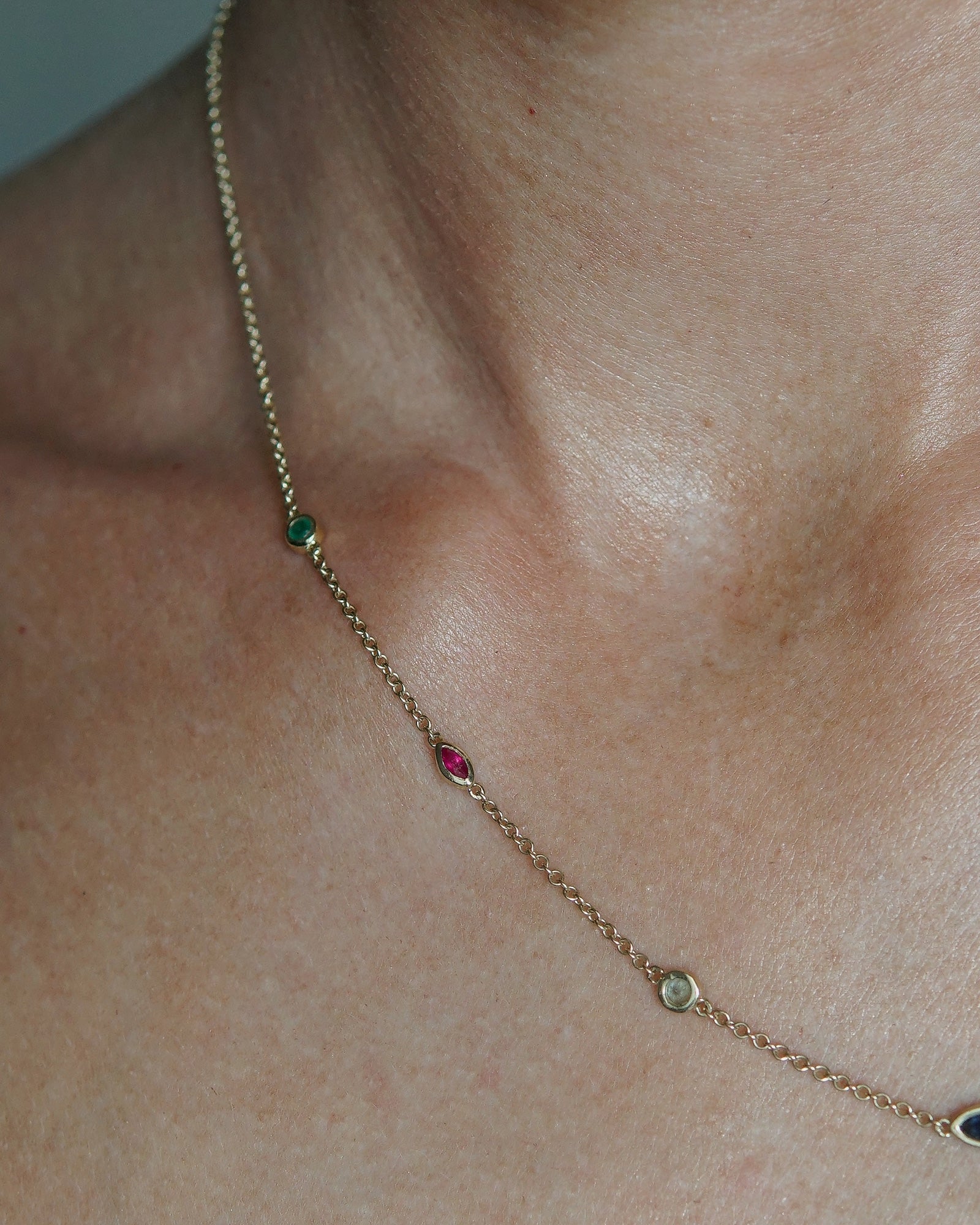 A delicate gold chain necklace featuring multiple small, colorful gemstone settings along the chain, including green round -cut emerald, red ruby marquise-cut stones, blue round -cut tourmaline, and d blue sapphire marquise-cut stones