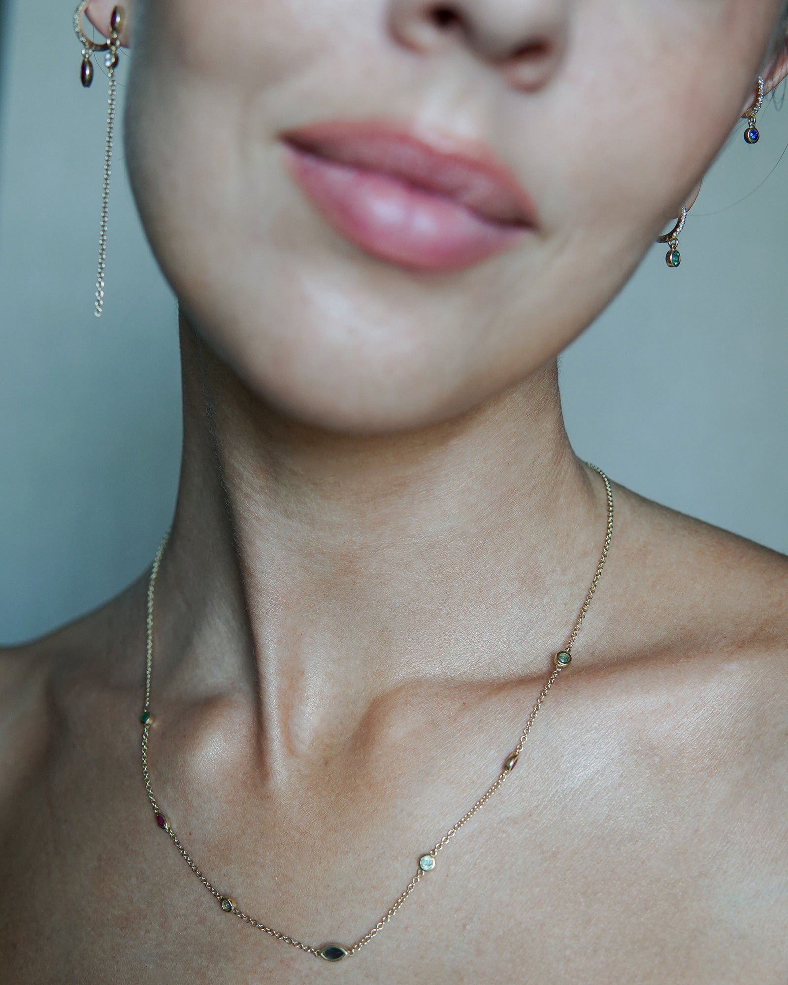 A delicate gold chain necklace featuring multiple small, colorful gemstone settings along the chain, including green round -cut emerald, red ruby marquise-cut stones, blue round -cut tourmaline, and d blue sapphire marquise-cut stones, displayed on a a woman's neck 