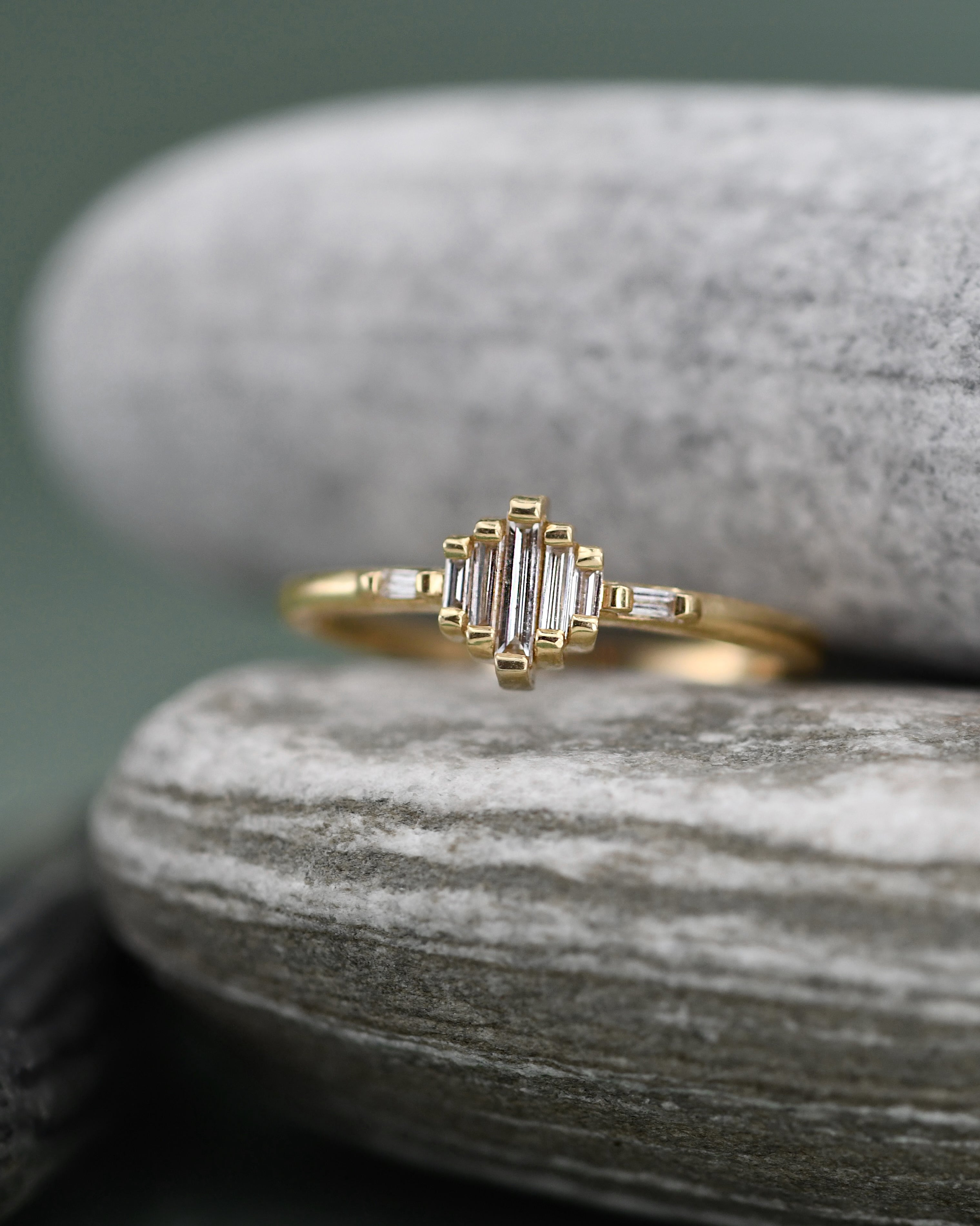 A gold ring with baguette and round-cut clear DIAMONDS, displayed on stacked gray stones against a soft green background.