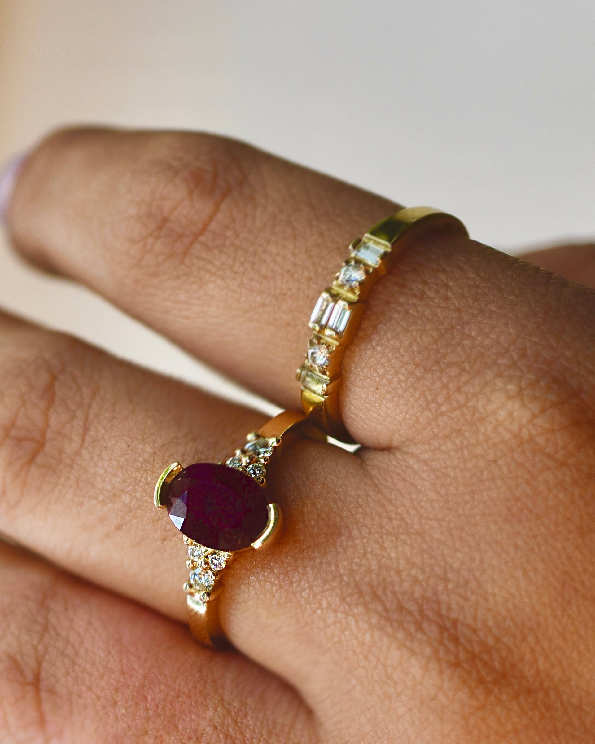 A close-up of a hand wearing two gold rings. The foreground ring features a large oval deep red ruby gemstone accented by small clear white diamonds, while the background ring is a delicate gold band with a row of clear baguette and round-cut white diamonds.
