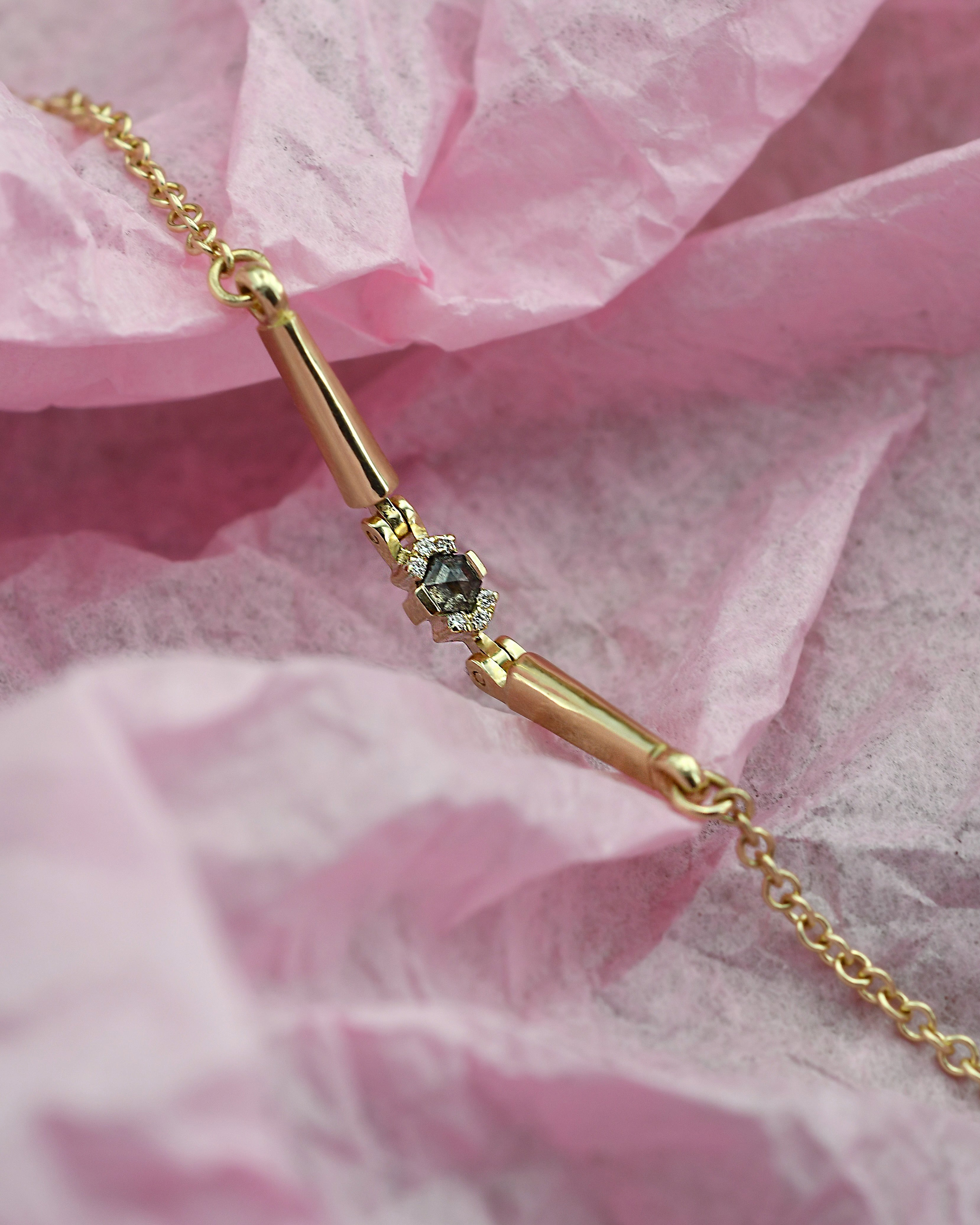 A gold bar bracelet with a central 
grey diamond, displayed on a crumpled pink paper background.