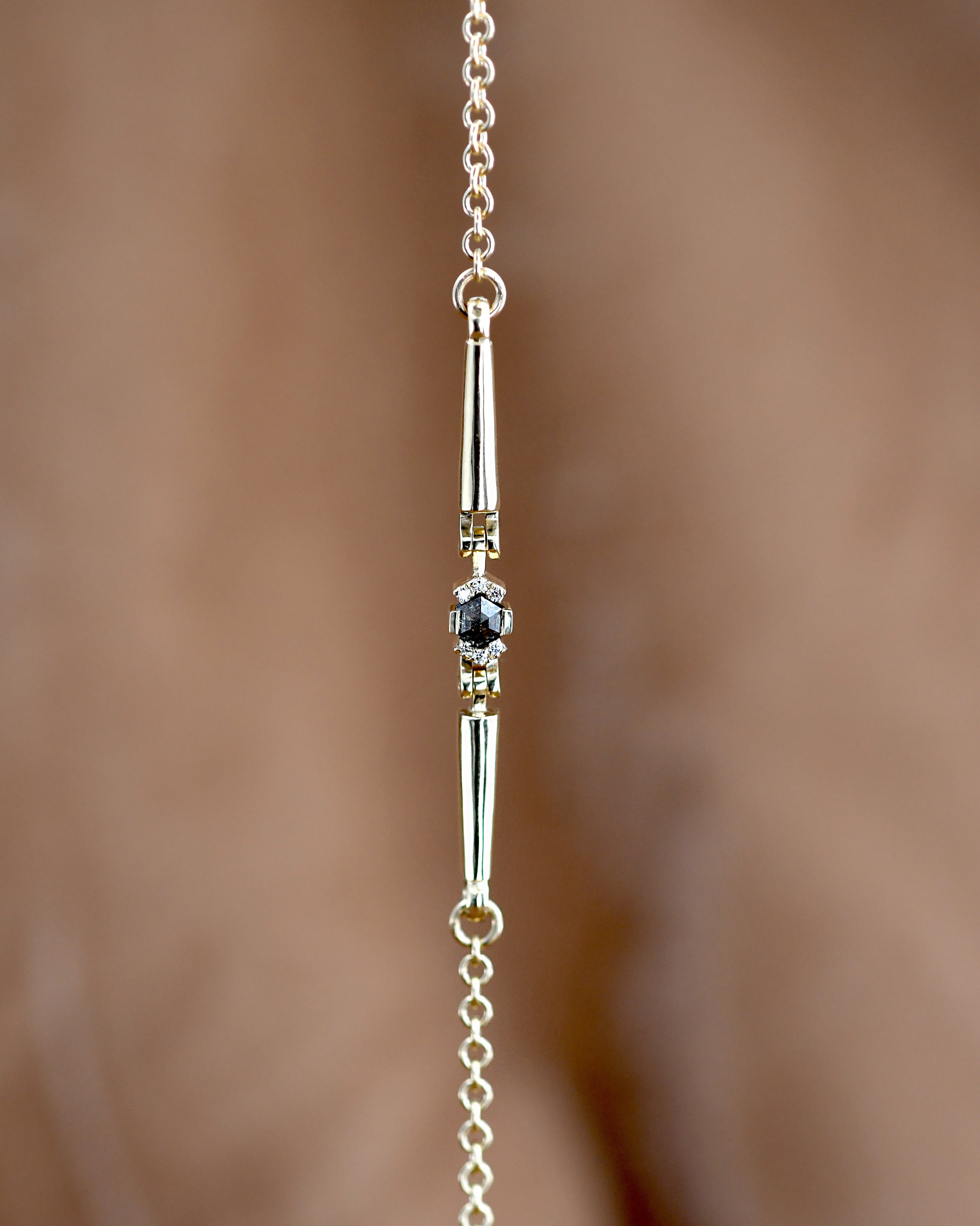 A close-up of a delicate gold brecelet featuring a central bar pendant with  faceted dark gray diamond, set against a soft, blurred warm brown background.