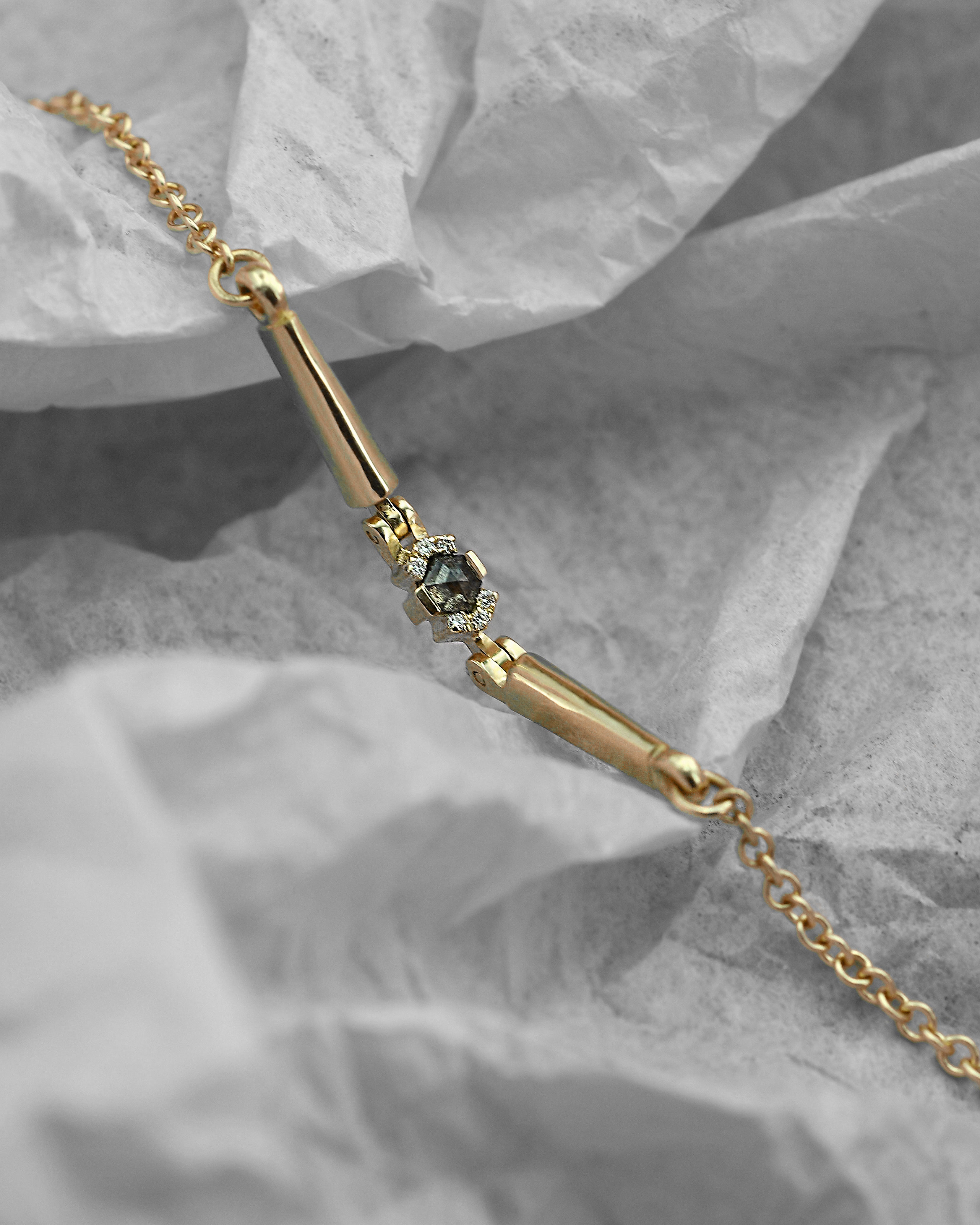 A gold bar bracelet with a central 
grey diamond, displayed on a crumpled gray paper background.