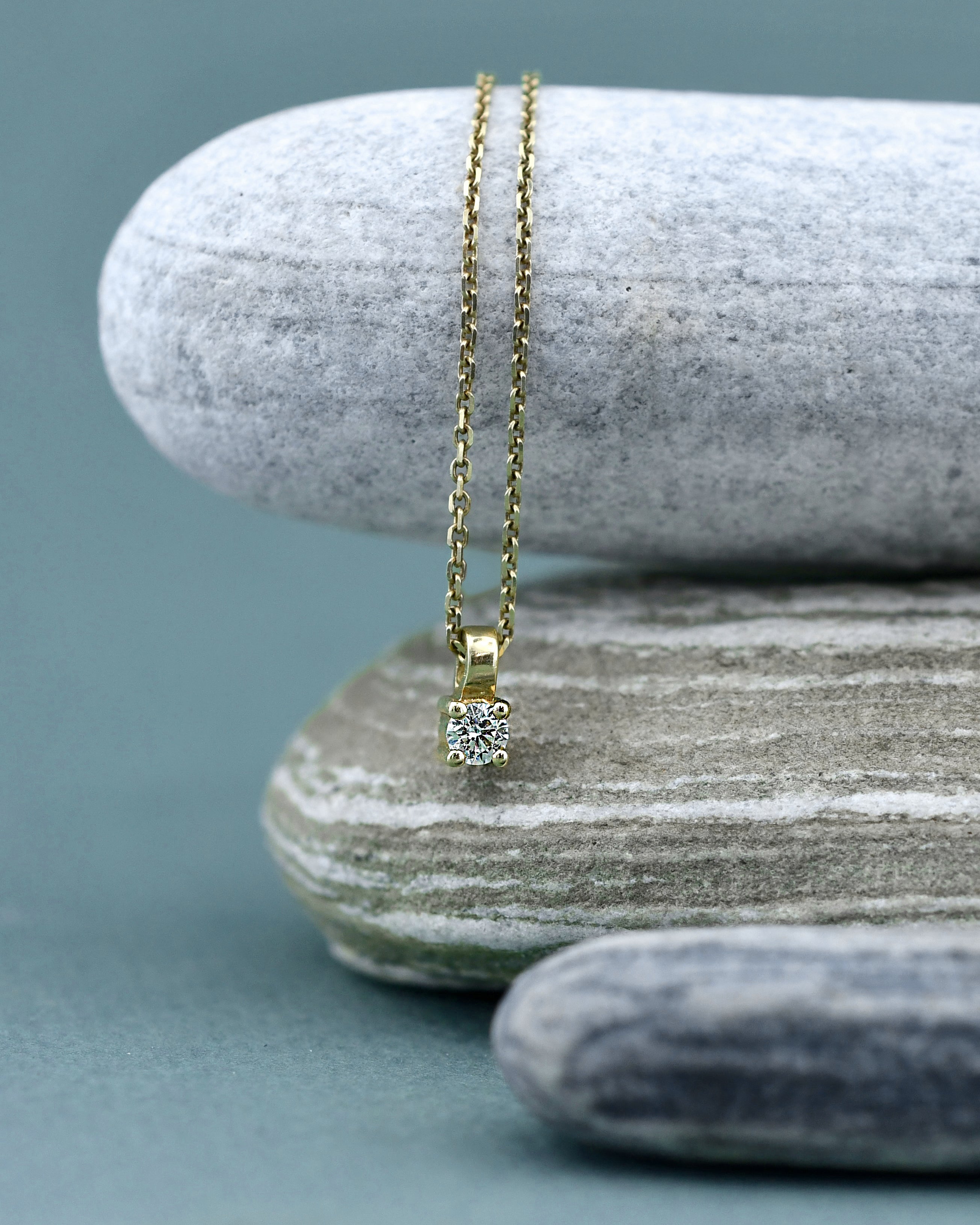 A delicate yellow gold necklace with a small square bezel-set clear diamond pendant, displayed on stacked gray stones against a muted blue-gray background.