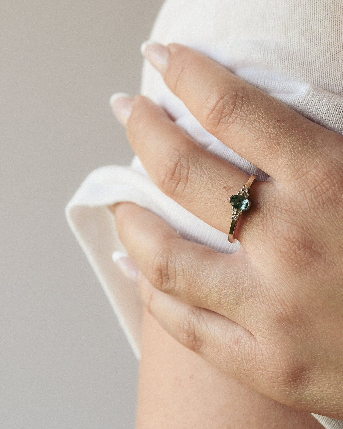 A close-up of a hand wearing a delicate gold ring with a green gemstone and white diamonds, resting on a soft white sleeve against a light neutral background.