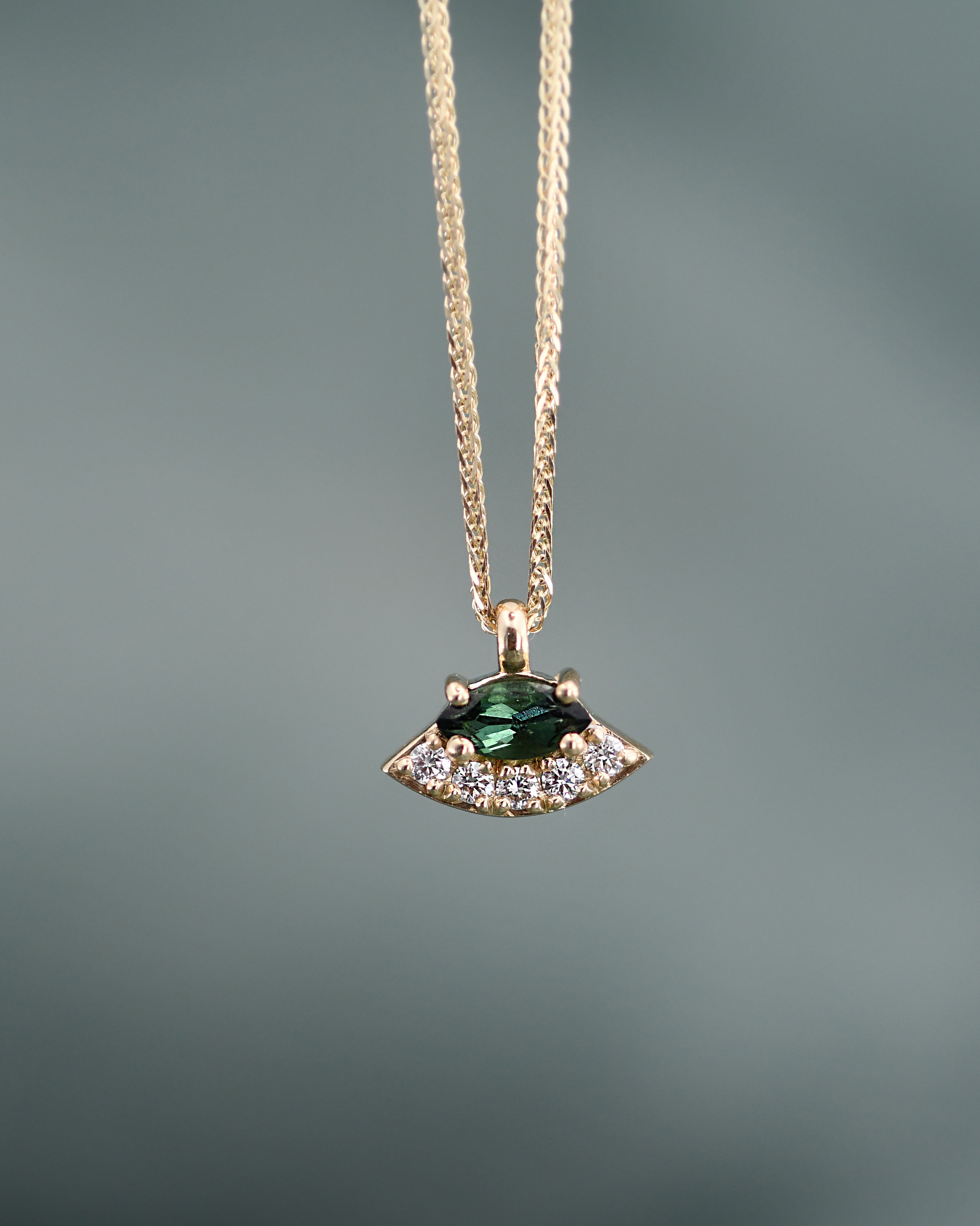 A gold chain necklace with a pendant featuring a marquise-cut green gemstone surrounded by small clear diamonds, shown against a soft gray background.