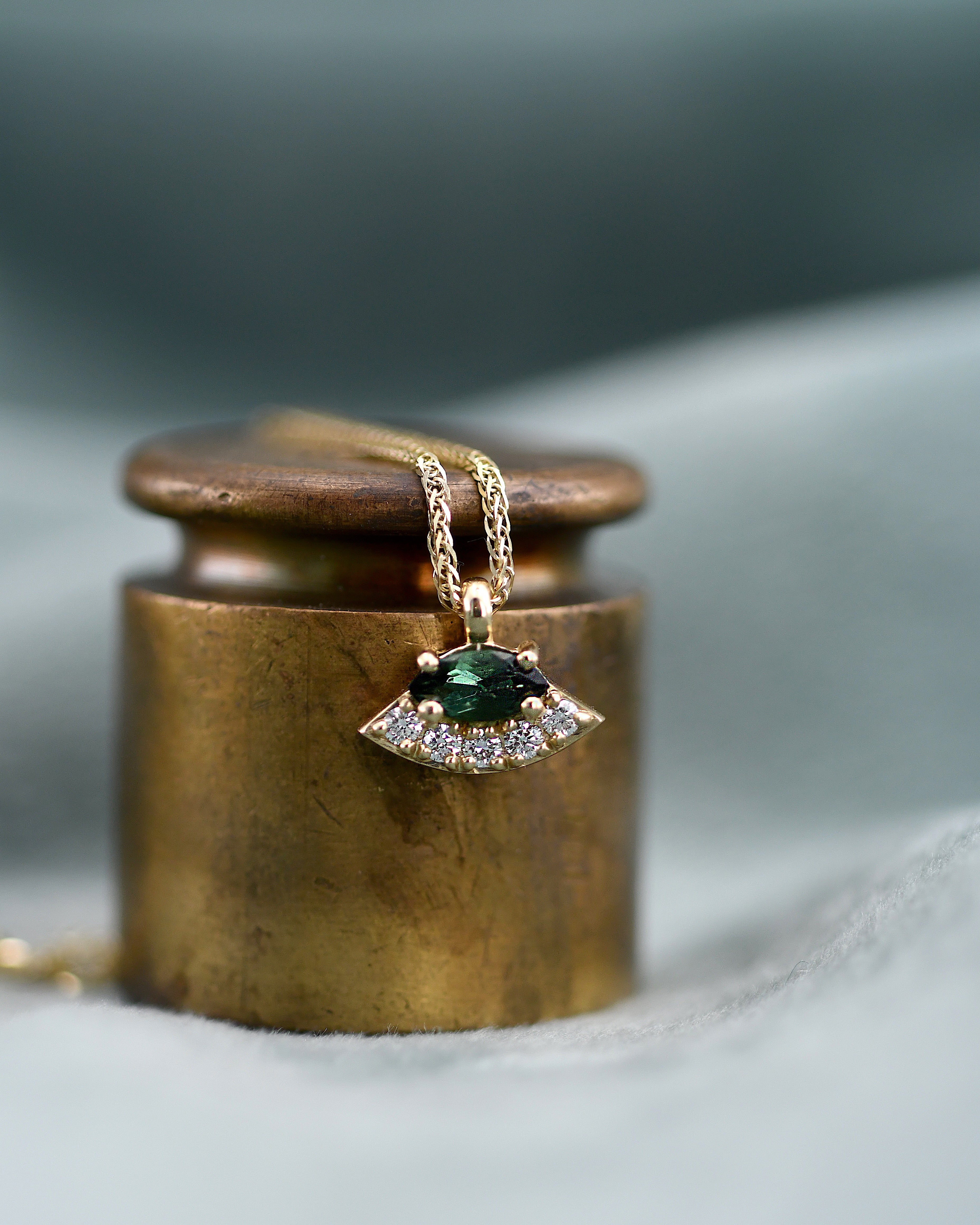 A gold pendant necklace with a green oval gemstone and small clear diamonds, displayed on a textured gold cylindrical stand against a soft gray background.